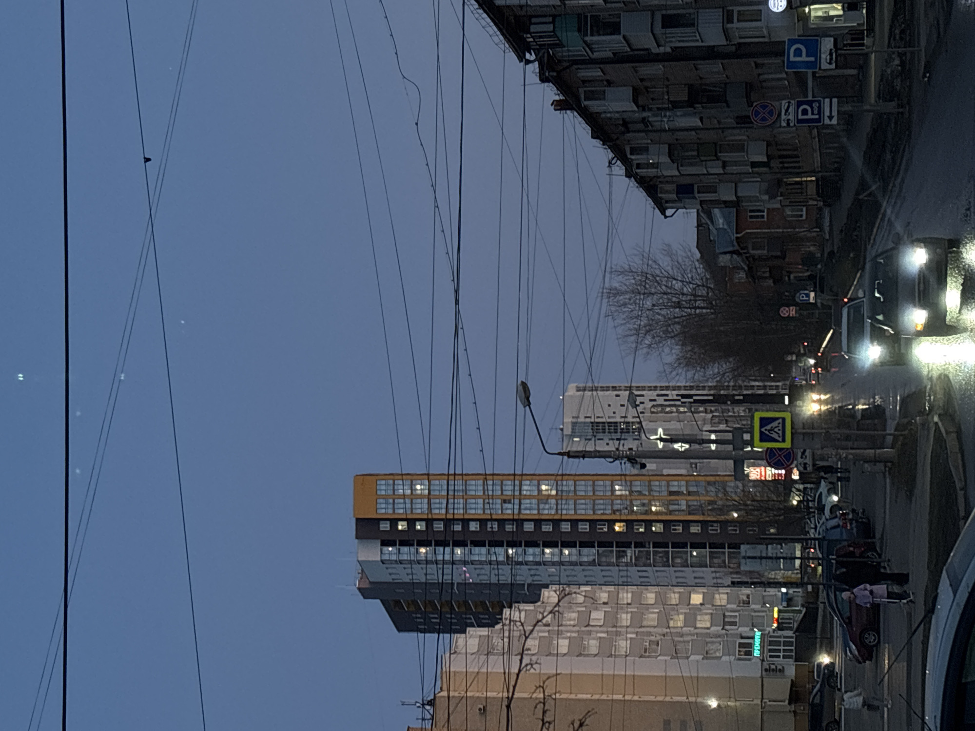 The image depicts a street scene in Perm, Russia, at dusk. Several multi-story buildings are visible. Power lines crisscross the sky. A road is present with cars and pedestrians. Street lights illuminate the scene. Road signs are visible. One person is possibly entering a car.