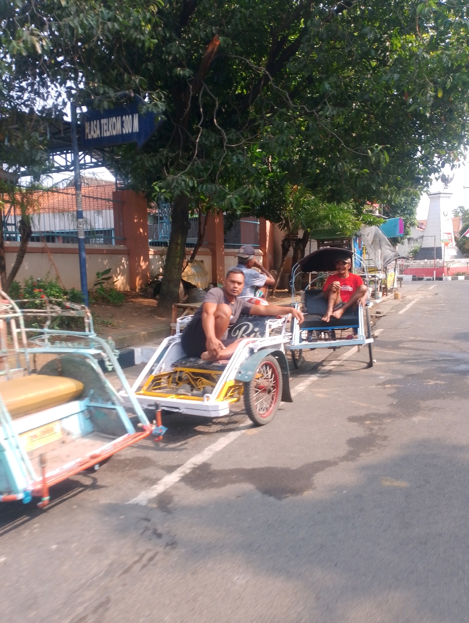 The image depicts a street scene in Probolinggo, Indonesia, featuring three pedicabs and several people. In the foreground, a light blue and yellow pedicab is partially visible on the left, with its passenger seat and wheel clearly in frame. To the right, another pedicab, white and yellow with a blue canopy, is occupied by two men. The man in the rear is wearing a red t-shirt and appears to be looking forward. The man in the front, wearing a dark t-shirt and dark shorts, is sitting with his legs extended and is looking towards the camera. A third pedicab is further in the background, also with a passenger. The street is paved and appears to be a road with a white line marking the edge. Trees with green foliage line the street, and a blue sign with white text reading "PLASA TELKOM 300 M" is visible on the left, indicating a direction and distance to a Telekom Plaza. Buildings with orange roofs are visible behind the trees. The overall lighting suggests it is daytime, possibly morning or afternoon, with sunlight casting shadows.