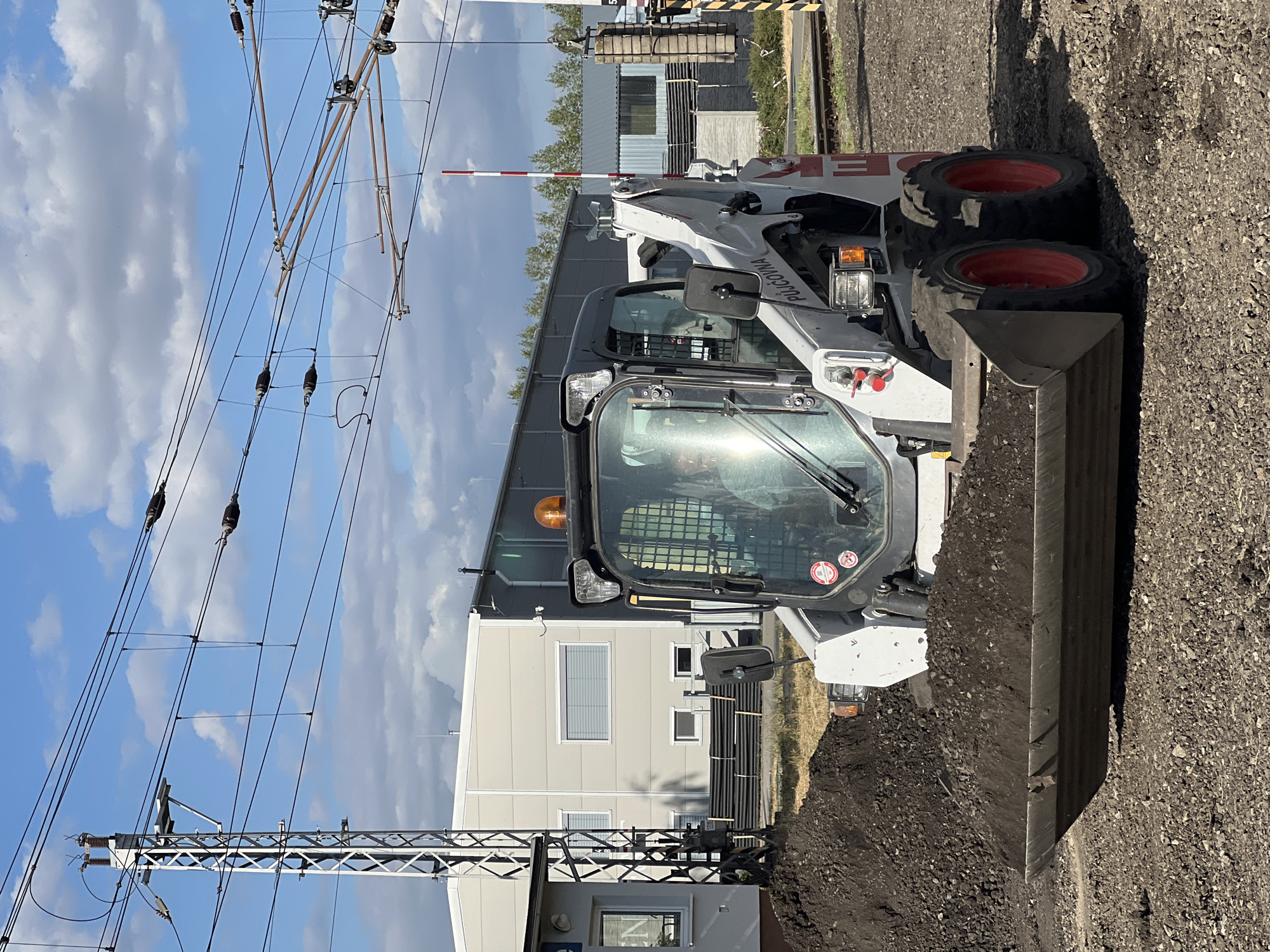 In Újezd u Brna, Czechia, a Bobcat compact track loader, identifiable by the text "PŮJČOVNA" (rental) on its side, is actively moving a pile of dark soil. The loader's bucket is filled with the soil, suggesting an ongoing construction or landscaping project. In the background, a light gray industrial building with several windows is visible, along with railway tracks and overhead power lines, indicative of the area's infrastructure. A red and white striped barrier arm, commonly seen at railway crossings, is also present. The sky is partly cloudy, suggesting a daytime scene. There is a person visible inside the cab of the loader.