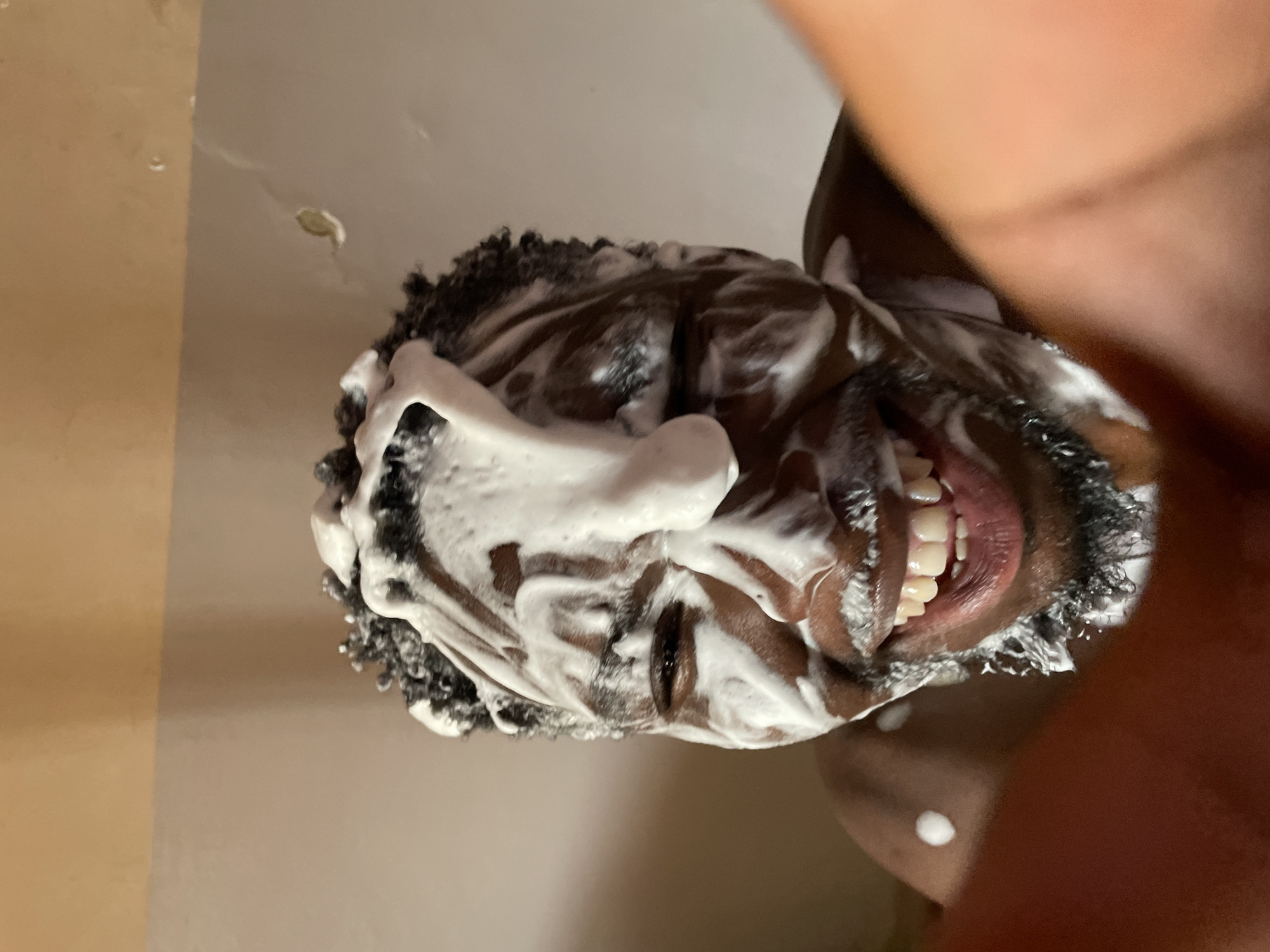An overhead close-up shot, rotated ninety degrees clockwise, features a person with dark skin and short, dark, curly hair. Their face and hair are thoroughly covered in white foam or suds, suggesting they are in the process of washing. The person is smiling broadly, revealing white teeth, which conveys a sense of happiness or playfulness. One eye is partially visible through the foam on the left side of the frame, looking towards the viewer. The background appears to be a light-colored, possibly tiled or painted, indoor surface, likely a bathroom wall or shower enclosure, with some water droplets visible. A blurred, dark-skinned arm or shoulder is visible on the right side of the image. No text is visible.