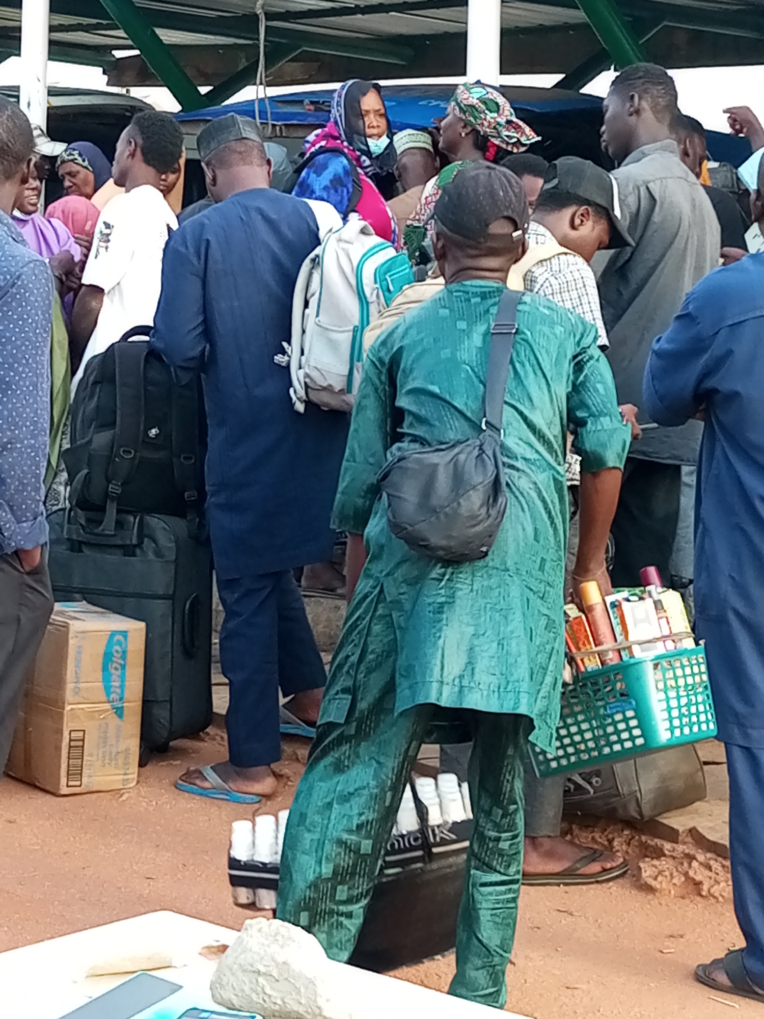 The image depicts a crowded outdoor scene, likely a transportation hub or market, in Gombe, Nigeria. Several individuals are visible, mostly men, dressed in traditional attire and head coverings. They are carrying luggage, including backpacks and suitcases, suggesting travel. In the foreground, a man in a green kaftan is holding a green basket filled with various items, possibly small goods for sale or personal belongings. Another man to his right is wearing a blue kaftan. In the background, a woman in a pink and blue headscarf is visible, along with other people and structures that suggest an open-air environment. The ground appears to be dirt or gravel. The overall impression is one of bustling activity and movement.