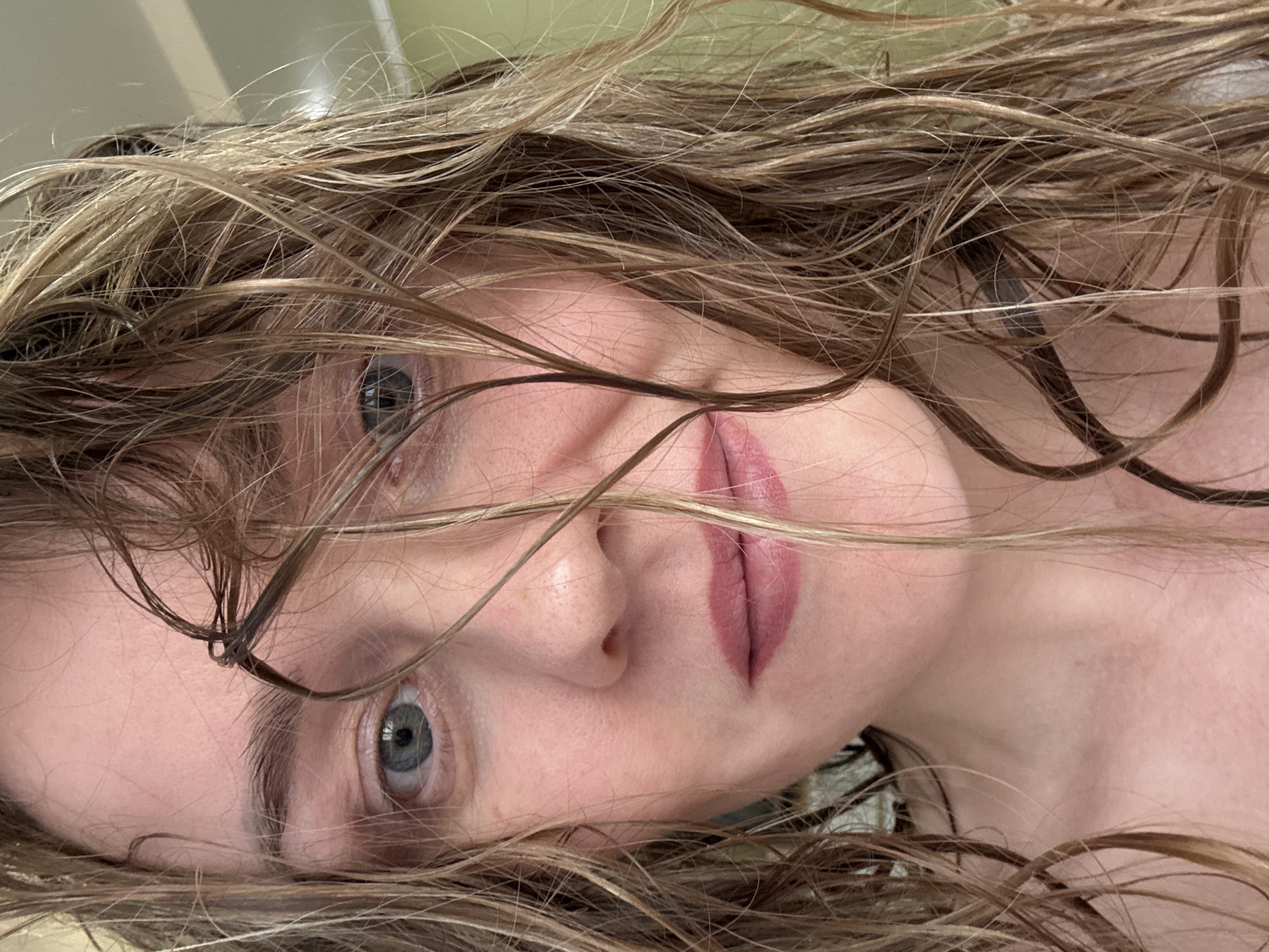 A close-up, intimate portrait captures a person, likely female, lying down and looking directly at the camera. The image is oriented with the subject's head rotated approximately 90 degrees clockwise, creating an unconventional and personal perspective. Her light brown or blonde hair, appearing wet and styled in loose, wavy strands, falls across her face, partially obscuring her forehead and cheeks.
The subject's blue-grey eyes are open and gaze intently towards the viewer. Her lips, enhanced with a soft pink or mauve lipstick, are full and slightly parted. Her fair skin displays natural texture with visible pores, especially around the nose and upper lip, emphasizing the close proximity of the shot. A portion of her bare shoulder and neck is visible in the lower right, suggesting a relaxed or casual attire.
The background is softly blurred, showing indistinct light tones, possibly a ceiling or wall in an indoor setting. The lighting is even and soft, highlighting the subject's features without harsh shadows. The overall impression conveys a moment of quiet introspection or a styled shot emphasizing natural, wet-hair beauty. There is no visible text in the image. The scene offers no specific location cues beyond being indoors, potentially in a private space in Chaikovsky, Russia.