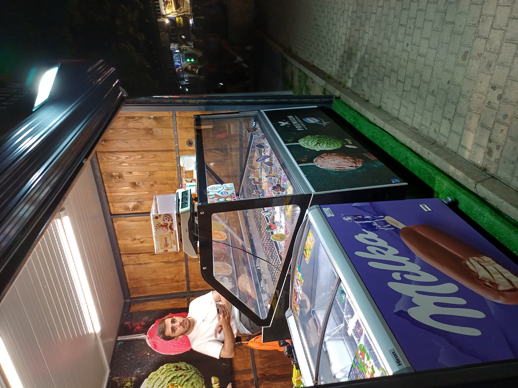A young man wearing a white t-shirt is standing behind an ice cream display freezer and counter in an outdoor kiosk at night. He is looking at his mobile phone. The kiosk appears to be on a paved street, with a few people visible in the background in front of a pharmacy and ATM. The ice cream display has several flavors visible behind a glass panel, and there is a large freezer with a "Milka" advertisement featuring a cow and an ice cream bar. The kiosk is illuminated by overhead lights, and the surrounding area is dimly lit. The location is Varna, Bulgaria.