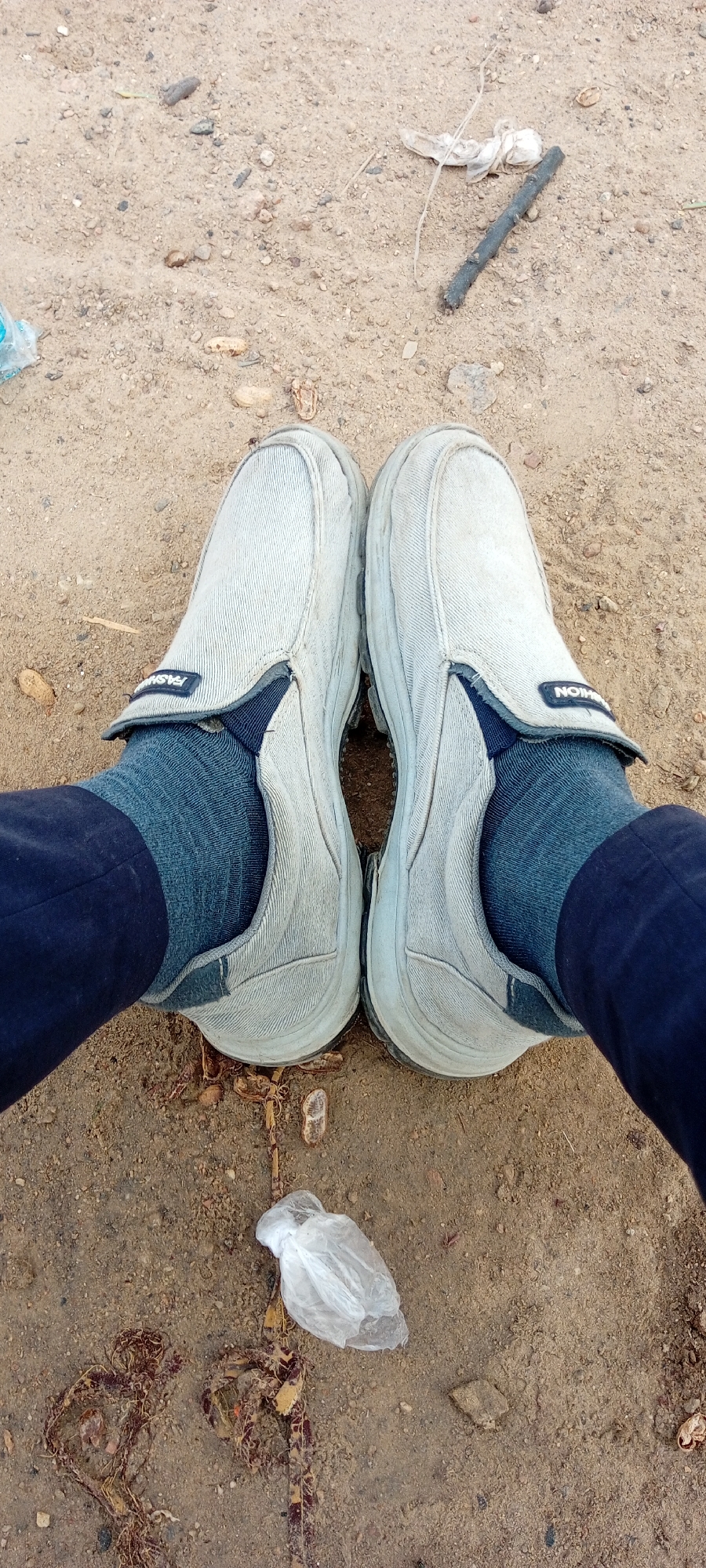In Kano, Nigeria, this image captures a close-up, ground-level perspective of a person's feet wearing light-colored canvas slip-on shoes and dark blue socks. The shoes are slightly dirty, and a dark blue fabric is visible at the ankles, suggesting dark pants are being worn. The shoes have a dark trim and a small black label with white lettering that reads "FASHION" on the right shoe. The ground beneath is composed of dry, sandy soil with scattered small pebbles and debris, including a dried plant stem, a dark stick, and a crumpled white plastic bag. The overall environment appears to be an outdoor, unpaved area, possibly a roadside or a less-maintained public space within Kano.