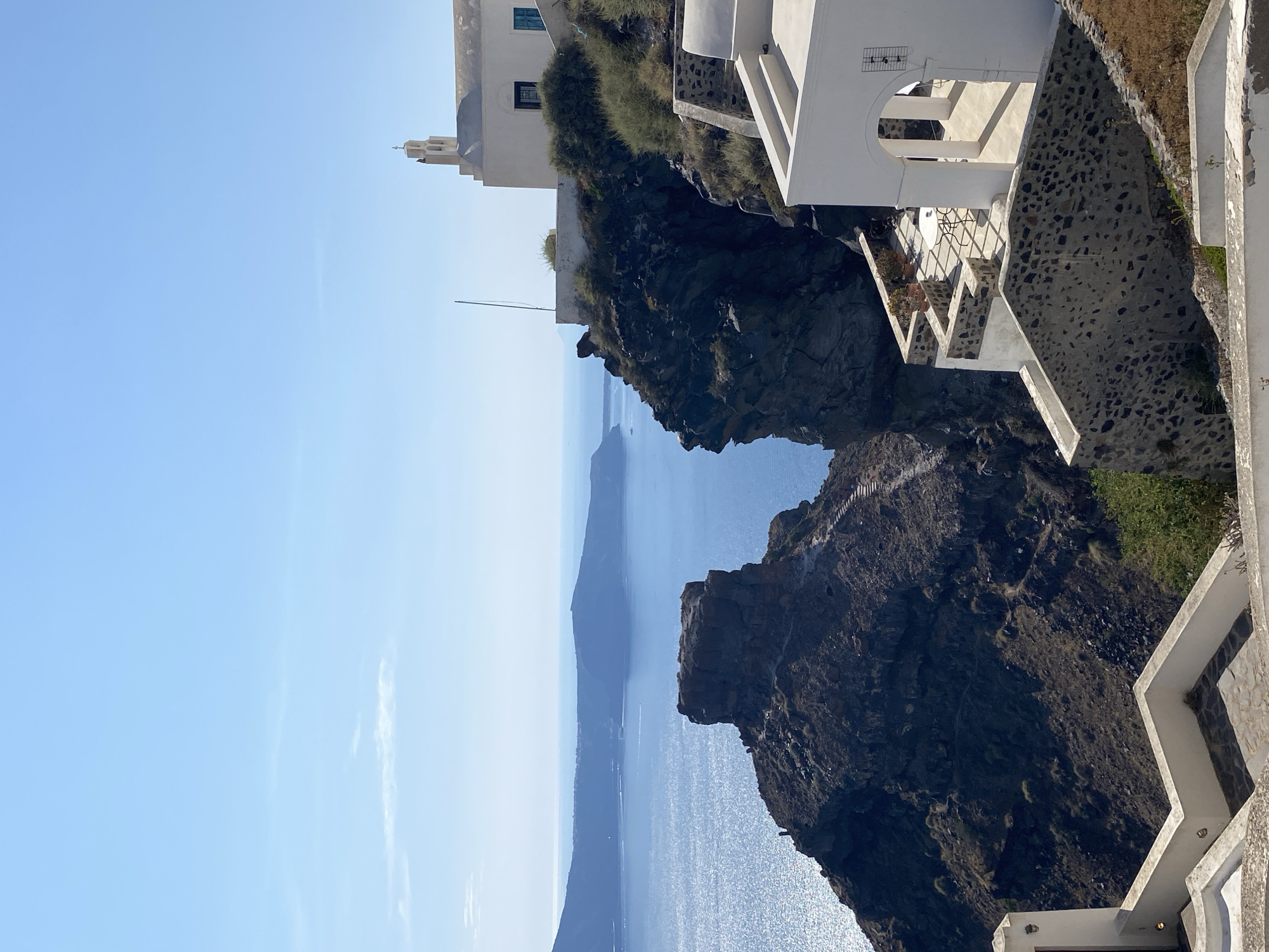 This is a vertical, eye-level, outdoor shot of a cliffside in Thira Municipal Unit, Greece, with the Aegean Sea in the background. The bright blue sky fills the top third of the image, with a few wispy clouds on the horizon. A white building with a bell tower and a cross is perched on the cliff to the right, overlooking the sea. In the foreground, white buildings with stone accents create a tiered effect, leading down towards the sea. The rocky cliff face is covered in sparse, dry vegetation. The sea itself is a deep blue, sparkling with sunlight, and a distant island chain is visible across the water.