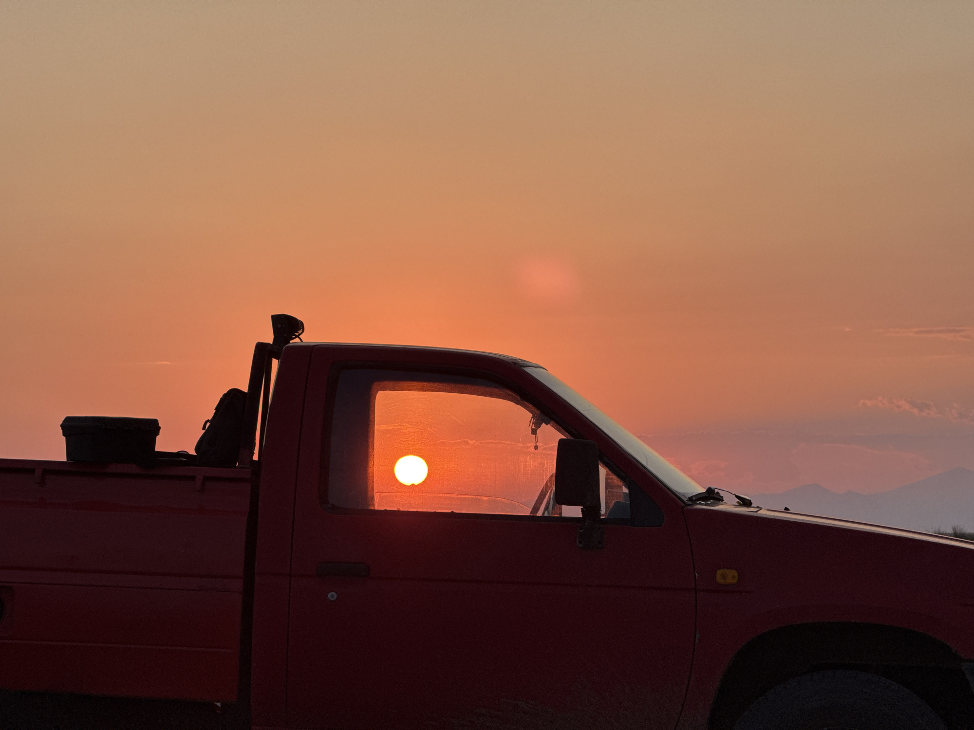 The image depicts a red pickup truck parked against a sunset sky. The sun, a bright orb, is visible through the truck's side window. A black toolbox and a backpack are placed in the truck bed. The truck is silhouetted against the warm orange and pink hues of the sunset. In the distance, faint outlines of mountains are visible. The scene appears to be set in an open area, possibly rural, and given the context, is located in Imeros, Greece. There are no people visible in the image.