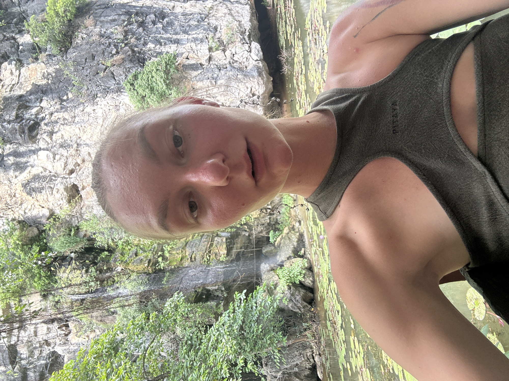 A young woman with fair skin and light brown hair pulled back poses for a selfie in a natural setting. She is wearing a dark green, cutout top. Her face is the primary focus, with her mouth slightly open. The background features a rocky cliff face with sparse vegetation, a cascading waterfall, and a body of water covered in lily pads. The location is identified as Nam Hoa Lư Ward, Vietnam.