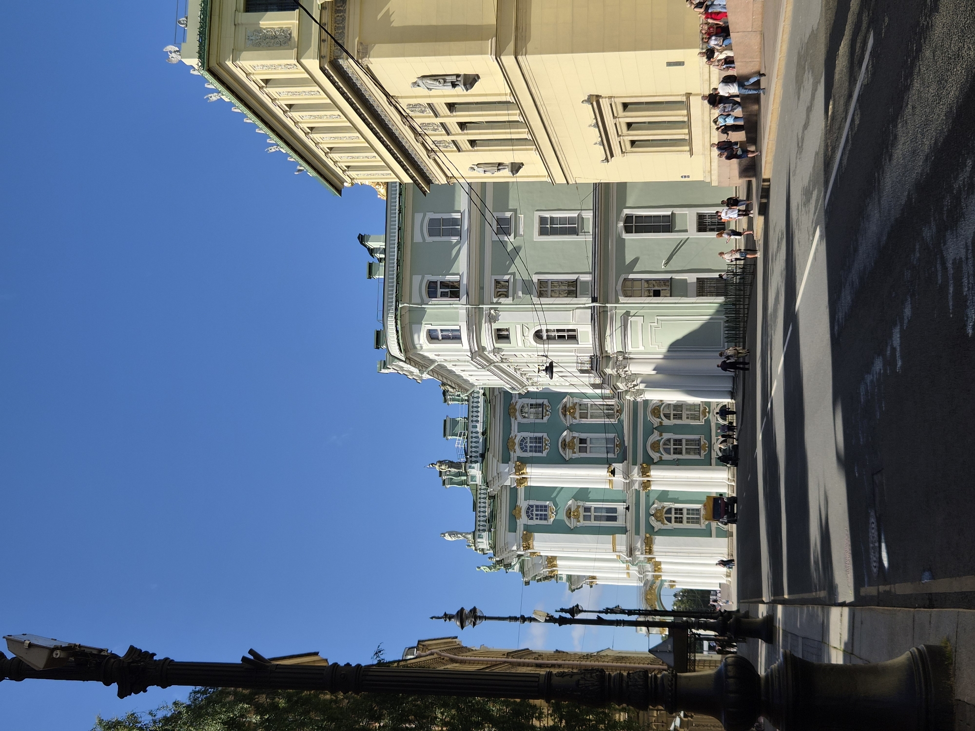 The image captures a street scene with grand historical buildings under a clear blue sky. On the left, a tall, ornate black lamppost stands prominently. To its right, a row of neoclassical buildings stretches into the distance, characterized by their light green and pale yellow facades, adorned with white columns, intricate moldings, and statues. Several people are visible on the sidewalk and the street, some walking, others standing or sitting on a raised stone ledge. A yellow truck and a black car are parked on the street. Sunlight casts long shadows across the asphalt, indicating it is daytime. The overall impression is one of architectural grandeur and a bustling urban environment.