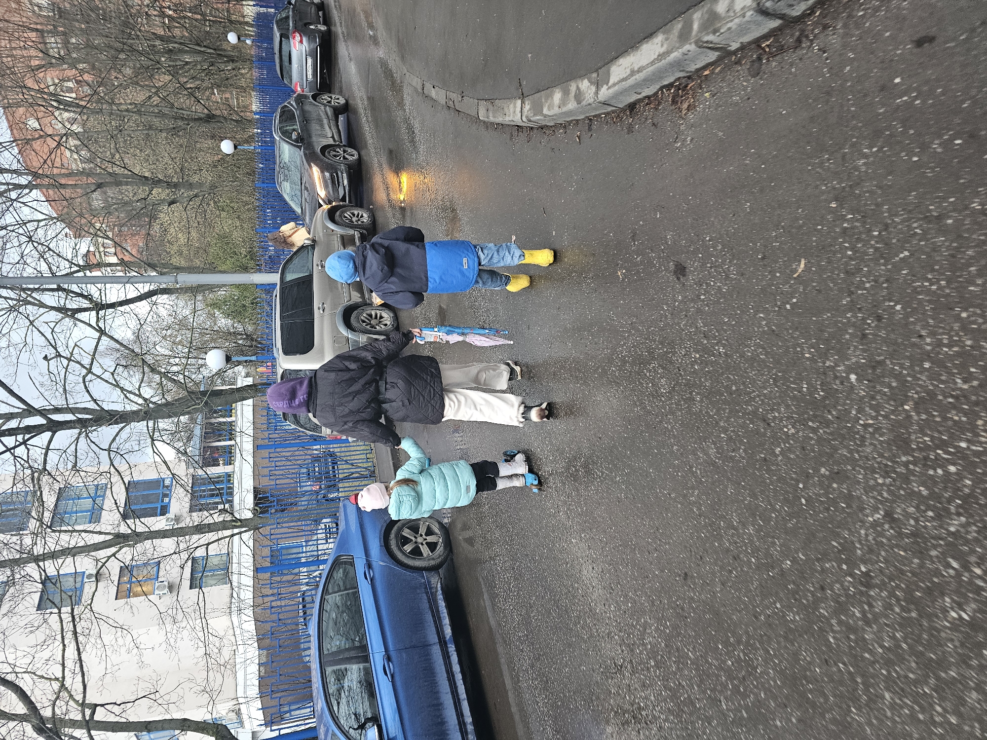 The image depicts three individuals walking along a wet street in what appears to be an urban environment. The location is identified as Khimki, Russia.
The group consists of an adult and two children. The first child, wearing a blue jacket and hat with yellow boots, is walking ahead. The adult is in the middle, wearing a black jacket, light pants, and a purple hat, holding an umbrella. The second child, wearing a light blue jacket and a pink hat, is lagging behind, possibly on some sort of wheeled footwear.
The scene is alongside a street lined with parked cars. A blue car is parked closest to the viewer, adjacent to a blue fence. Several other vehicles, including silver and black cars, are visible in the background. The area appears to be residential, with apartment buildings visible in the distance. Bare trees are present, suggesting it is late fall or early spring. The ground is wet, indicating recent or ongoing rain.