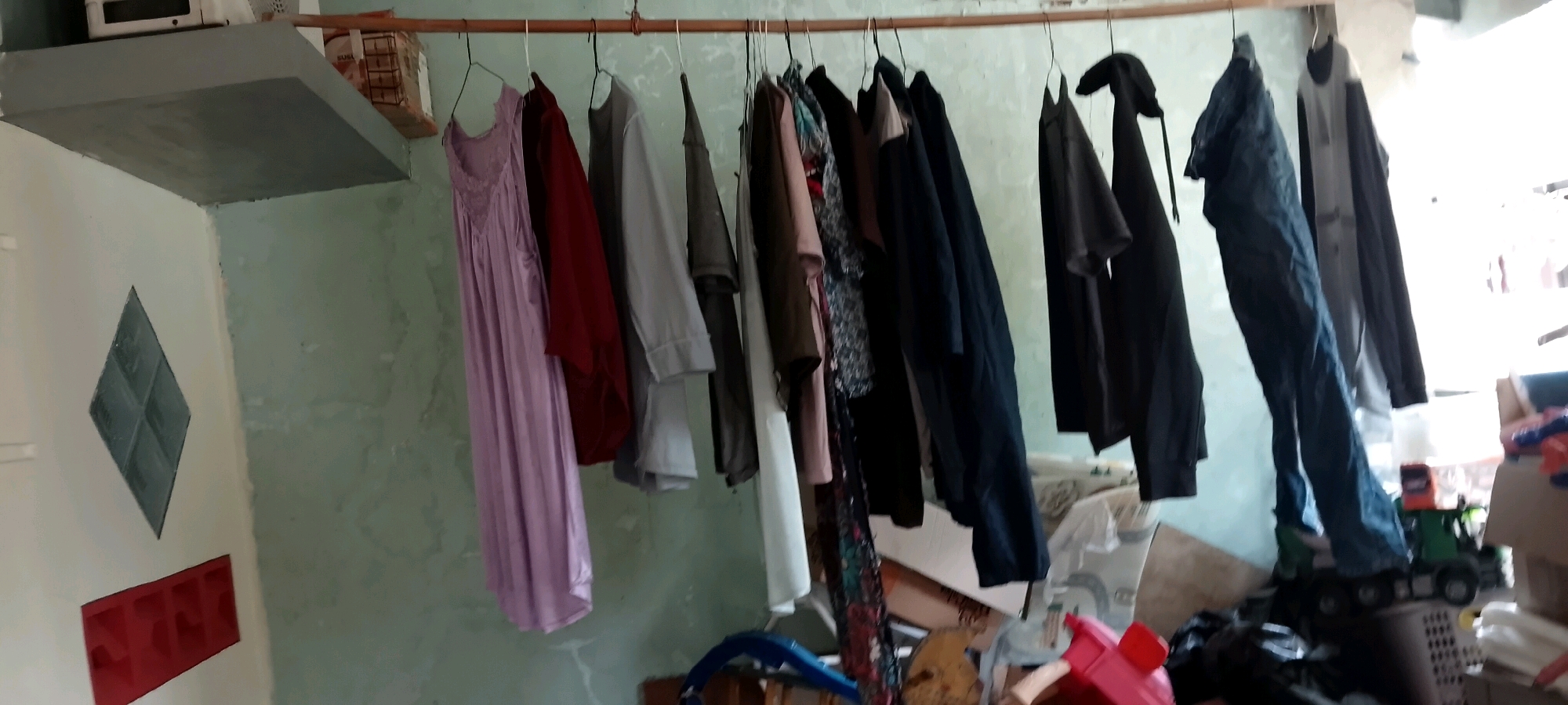 The image shows an indoor scene, likely a residence or workshop, in Satriamekar, Indonesia. A wooden clothesline is suspended horizontally across the frame, from which various garments are hanging. These include a long, light purple dress, a maroon garment, a grey shirt, a dark grey t-shirt, several dark-colored jackets or shirts, and a pair of blue jeans. To the left of the clothesline, a white wall features a diamond-shaped inset with dark green panes and a red rectangular object with multiple openings below it. A shelf is visible above the wall, supporting a white appliance and a cardboard box. The background consists of a pale green wall with visible imperfections. In the lower right portion of the image, a toy truck with a green cab and an orange and red trailer is partially visible, along with other clutter. The overall environment appears to be utilitarian and somewhat disorganized.