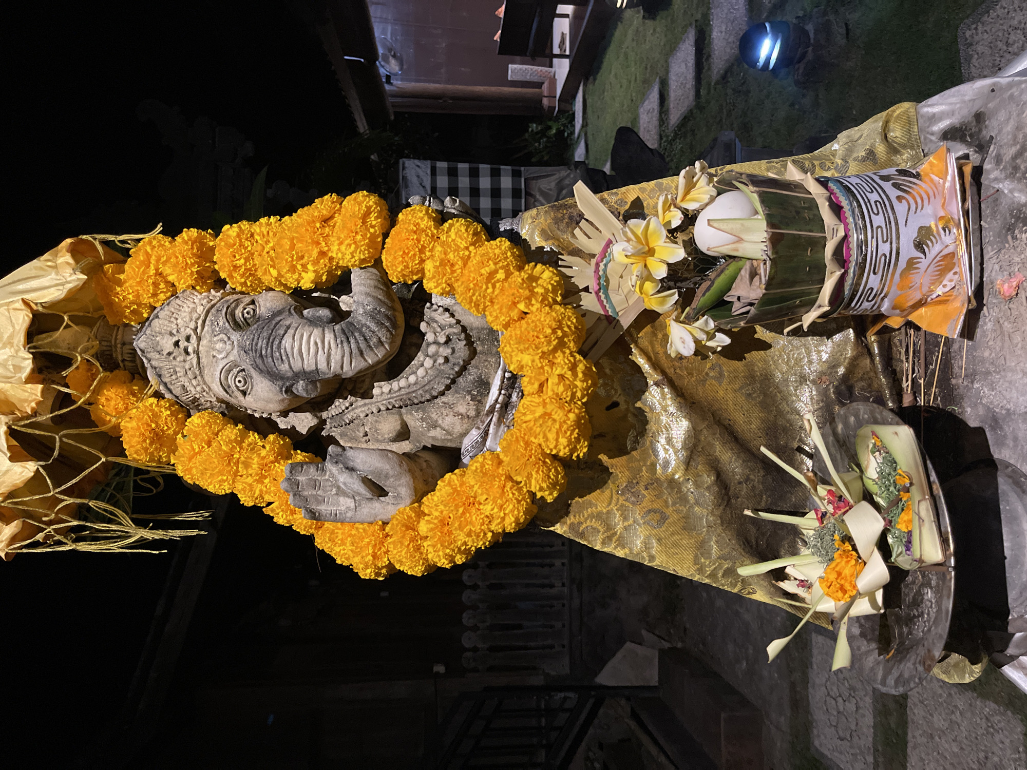 A stone statue of Ganesh, adorned with a lei of bright orange marigolds and a golden cloth, is centrally positioned in a dimly lit outdoor setting. The statue's trunk is raised, and its right hand is held up in a gesture of blessing. In front of the statue, two offerings are placed on a metal tray and a decorative base. The offerings consist of woven leaf baskets filled with flowers, greenery, and what appear to be small white eggs or offerings. A yellow fabric umbrella covers the top of the statue, and more marigolds are draped around its head and chest. The background shows a dark, possibly wooden structure, a checkered fabric, and a grassy area with stepping stones. A blue light illuminates the grass in the background. The overall scene suggests a religious or ceremonial context, possibly at night.