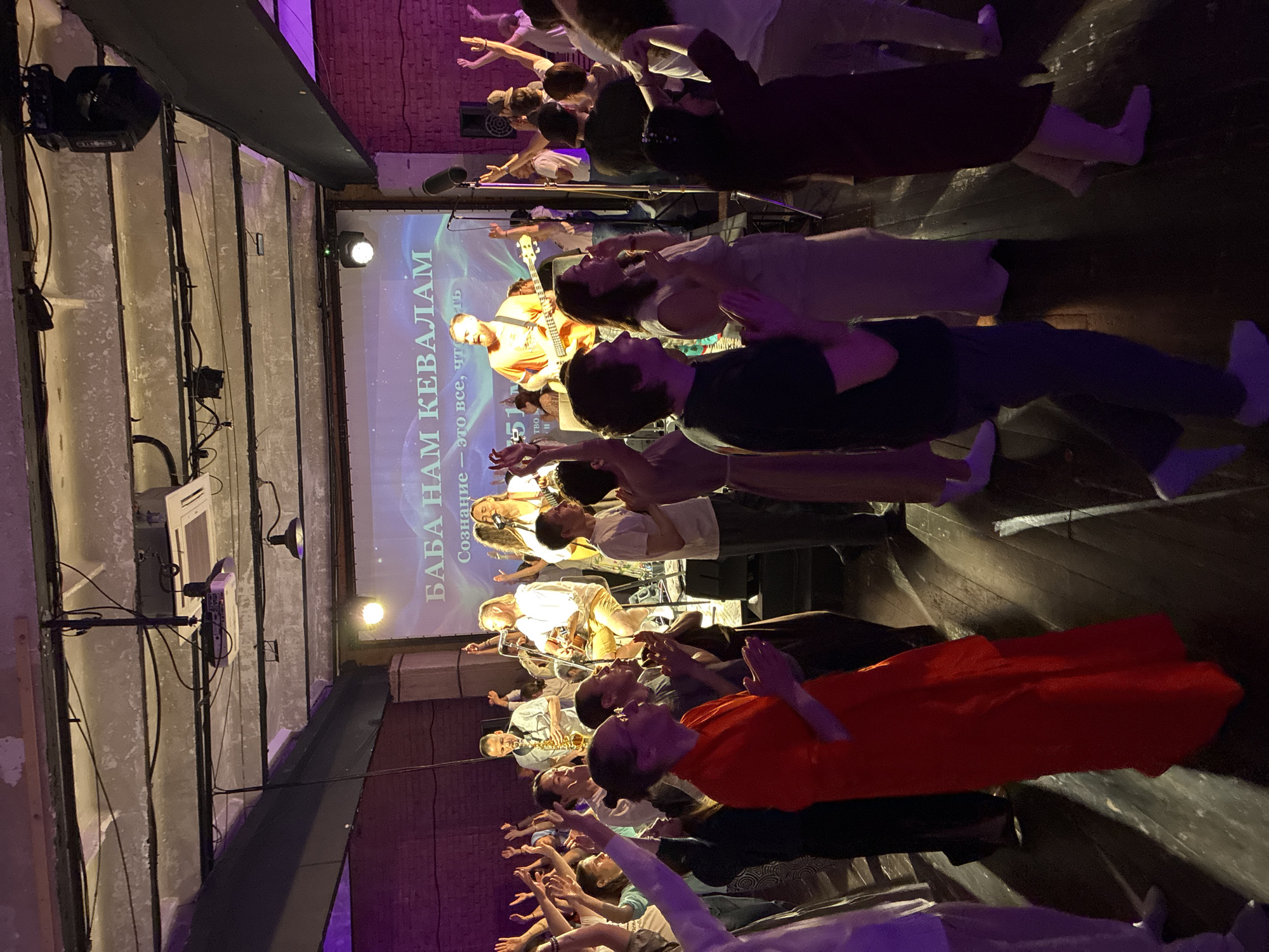 Here is a description of the image:
A group of people are attending a performance or gathering indoors. They stand facing a stage where musicians are performing. A screen behind the musicians displays text in Cyrillic script: "БАБА НАМ КЕВАЛАМ", and below that "Сознание - это все, что есть."
The attendees appear to be raising their arms in a gesture of participation or appreciation. The setting is a venue with exposed structural elements like metal beams and brick walls, possibly a renovated industrial space. The lighting is a mix of stage lights and ambient light with a purple hue.
While I can determine that this is likely in Moscow, Russia, due to the Cyrillic writing, it is difficult to pinpoint the exact location within the city without further information.