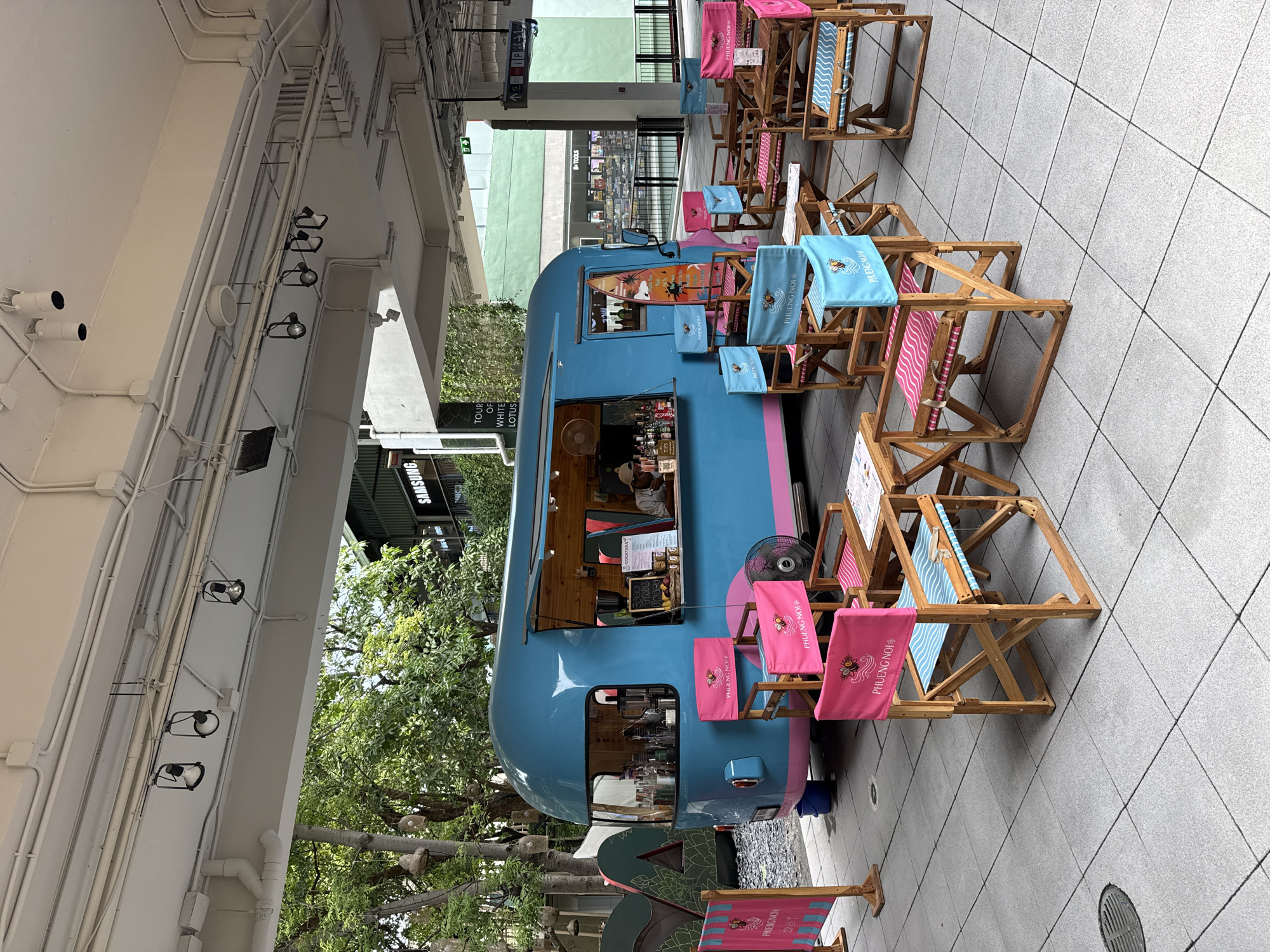The image depicts an outdoor seating area with a prominent blue and pink food truck serving drinks. The food truck is customized with a surfboard graphic on its side. Surrounding the food truck are several wooden chairs and tables, many of which are adorned with pink and blue fabric featuring the text "PHUENG NOI". A few pink banners with the same text are also visible. In the background, trees and buildings are present, suggesting an urban or semi-urban environment. The overall scene appears to be a commercial establishment, likely a cafe or bar, designed with a beachy or tropical theme. The location is the city of Ko Samui District, Thailand.
