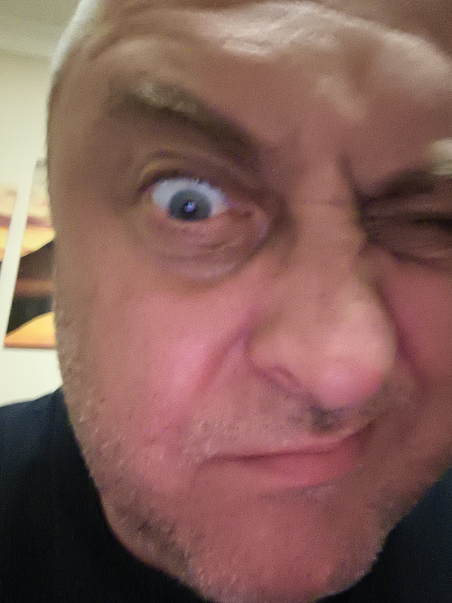 A tightly cropped, close-up portrait features the face of a middle-aged or older man, looking directly at the camera. His right eye (left side of the frame) is wide open with a blue iris, while his left eye (right side of the frame) is squinted or winking, creating a mischievous or playful expression. His mouth is closed, and his cheeks and chin show visible skin texture and light stubble. Wrinkles are evident on his forehead and around his eyes, consistent with his age.
His skin appears reddish, possibly due to lighting or emotion. Short, light-colored hair, which could be blonde or grey, is visible on the top and sides of his head. A dark-colored garment, likely a shirt or top, is visible on his lower left shoulder and neck area.
The background is very blurry and out of focus, suggesting an indoor environment. On the left side, abstract shapes in shades of yellow, orange, and dark brown are faintly discernible, possibly part of a piece of art or wall decor. The image exhibits noticeable digital noise or grain throughout, indicating it may have been taken in low light. No text is visible in the media file. The activity depicted is a man posing for a photo with a distinctive facial expression. The general location is Huntingdonshire, United Kingdom.