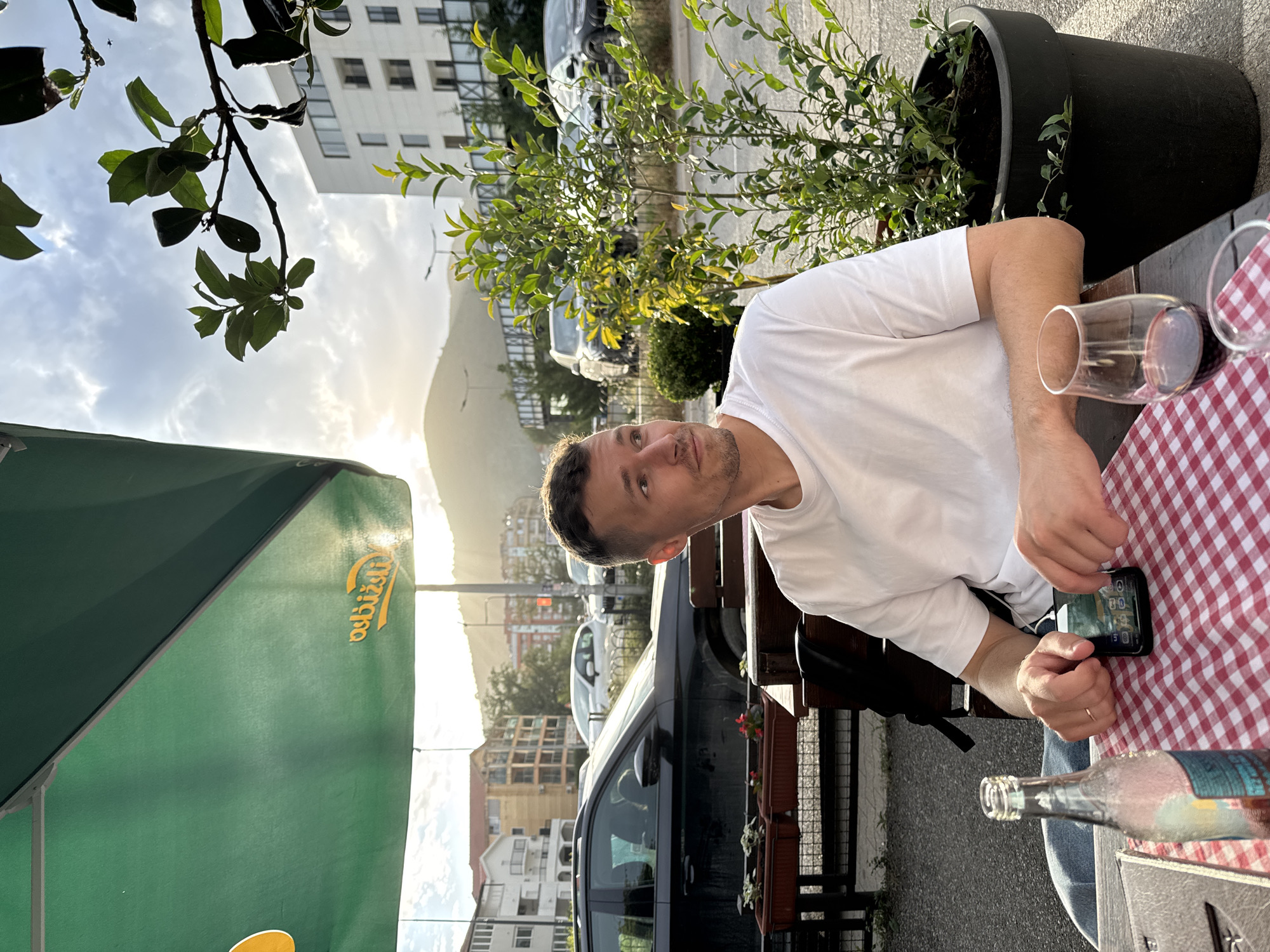 A photograph taken in Budva, Montenegro, shows a man seated at an outdoor table. He is wearing a white t-shirt and holding a black smartphone. A red and white checkered tablecloth covers the table, on which there is a wine glass, an empty glass bottle, and a potted plant. A green umbrella is partially visible on the left side of the frame. In the background, there are buildings, cars, and a mountain range illuminated by the setting sun.