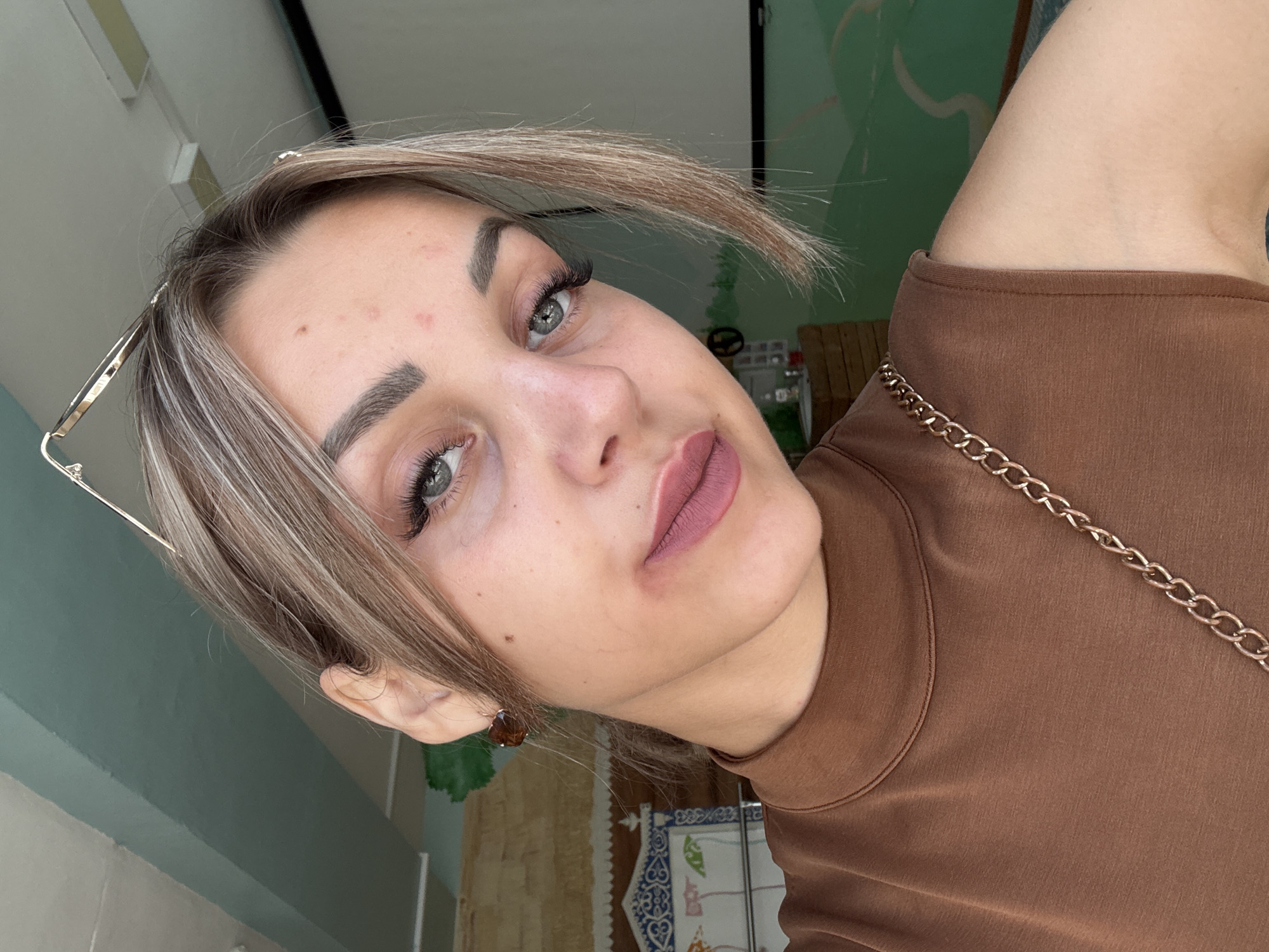 A young woman with light brown hair, styled in a casual updo with face-framing pieces, looks directly at the camera. She is wearing a brown, high-necked, sleeveless top with a gold chain strap draped across her shoulder. Her makeup includes winged eyeliner, false eyelashes, and matte pink lipstick. The background appears to be an indoor space with light green and white striped walls and a decorated panel on the left. The lighting suggests it is daytime. The woman's pose is a close-up selfie, angled slightly. The image does not contain any information or visual cues that would allow for a determination of its location in Yuzhno-Sakhalinsk, Russia, or any other specific urban area.