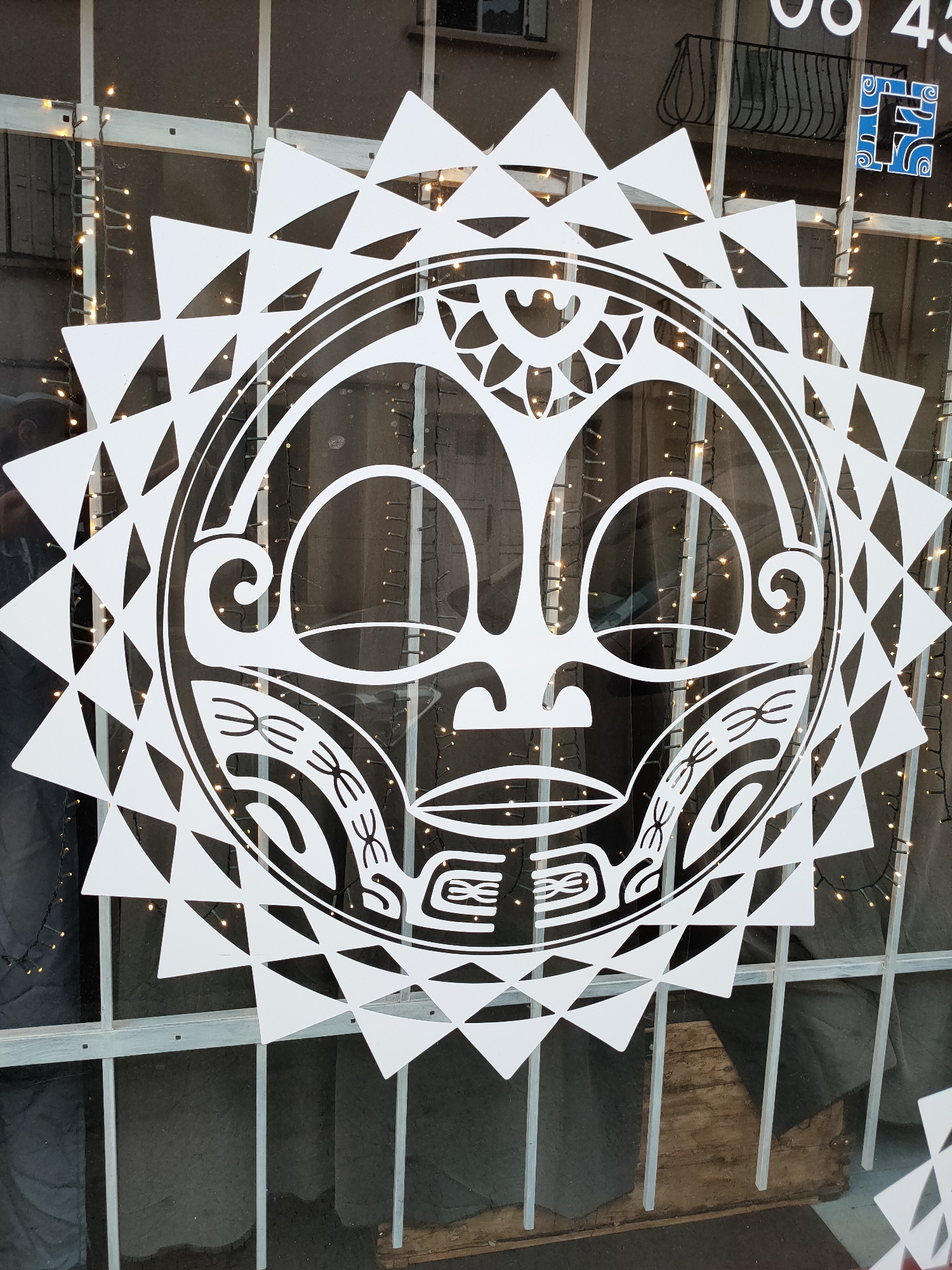 This is a close-up, eye-level, medium shot of a large white, sun-like graphic adhered to a storefront window. The graphic features a central face with stylized Polynesian-inspired motifs, surrounded by triangular points. Behind the graphic, a string of small, warm white fairy lights is visible, draped across a window frame and partially obscured by the graphic. The window itself has a light grey frame and vertical bars. In the upper right corner of the image, a blue and white logo with the letter "F" is partially visible, along with part of a dark numerical display that reads "08 43". A black wrought iron balcony is visible through the window in the background, as is a dark, indeterminate interior space. The image appears to be taken during daylight hours, possibly in the city of Perpignan, France.
