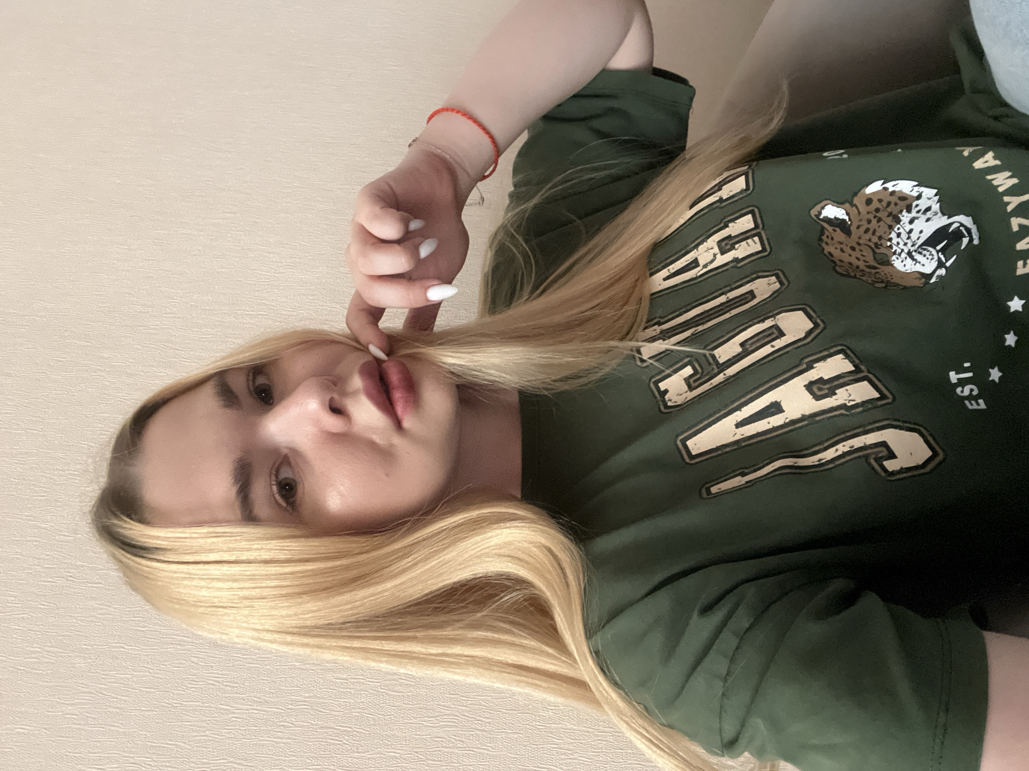 A young woman with long blonde hair, fair skin, and light brown eyes poses for a selfie. She is wearing a dark green t-shirt with a jaguar graphic and the word "JAGUAR" in large, distressed letters. Her left hand is raised to her lips, and she is wearing white nail polish and a red bracelet on her right wrist. The background is a plain, textured wall. The image quality is poor, with grainy resolution and unnatural lighting. No specific location in Krasnodar, Russia, can be identified.