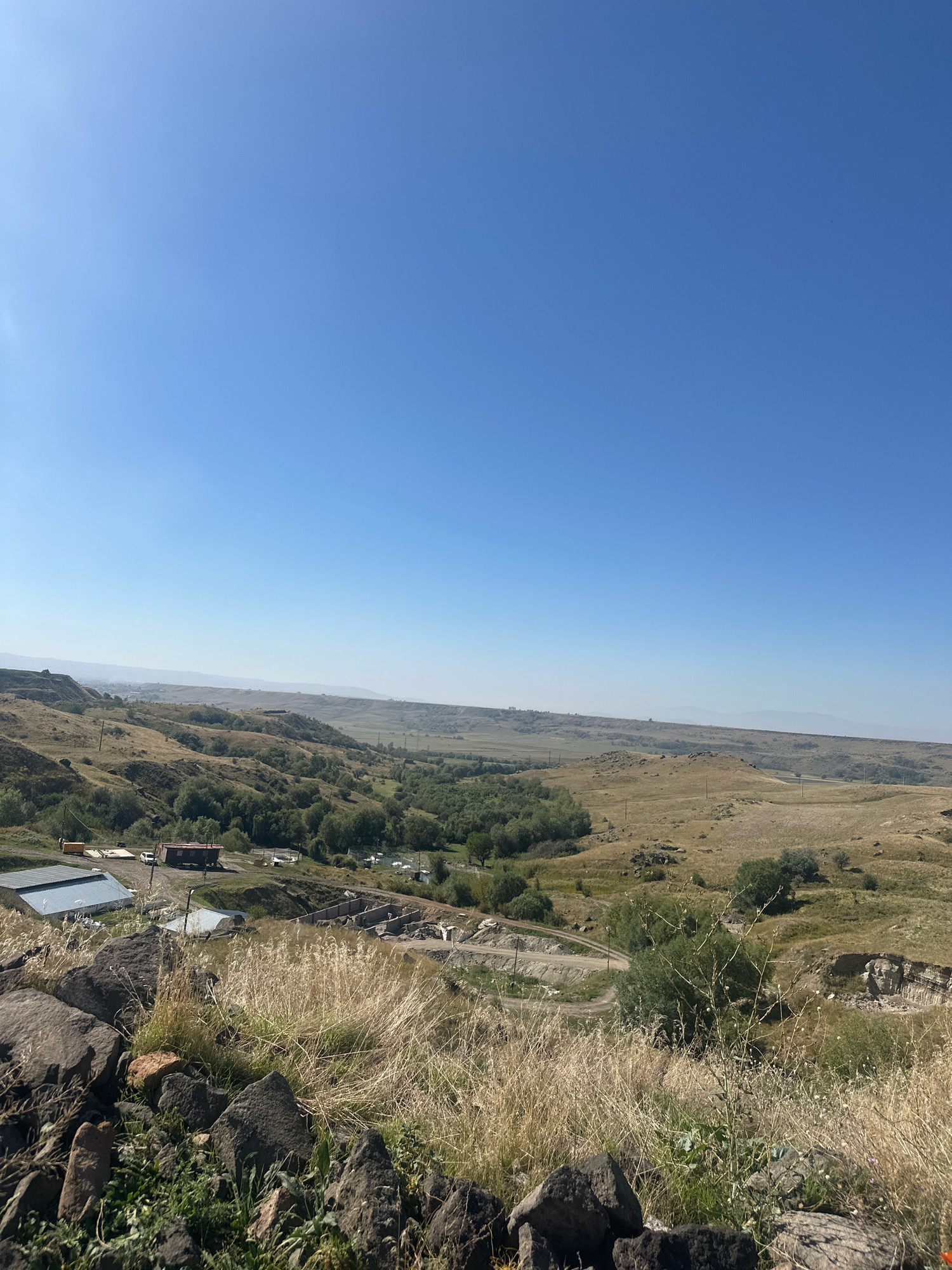 The image depicts a wide-angle view of a dry, hilly landscape under a clear blue sky. In the foreground, dark rocks and dry grass are prominent. In the midground, there are several structures, including buildings with corrugated metal roofs, a trailer, and a construction area with excavated earth and concrete foundations. Further in the distance, a valley with trees and a winding road can be seen. The background consists of rolling hills and a hazy horizon. The scene suggests a rural or semi-developed area.