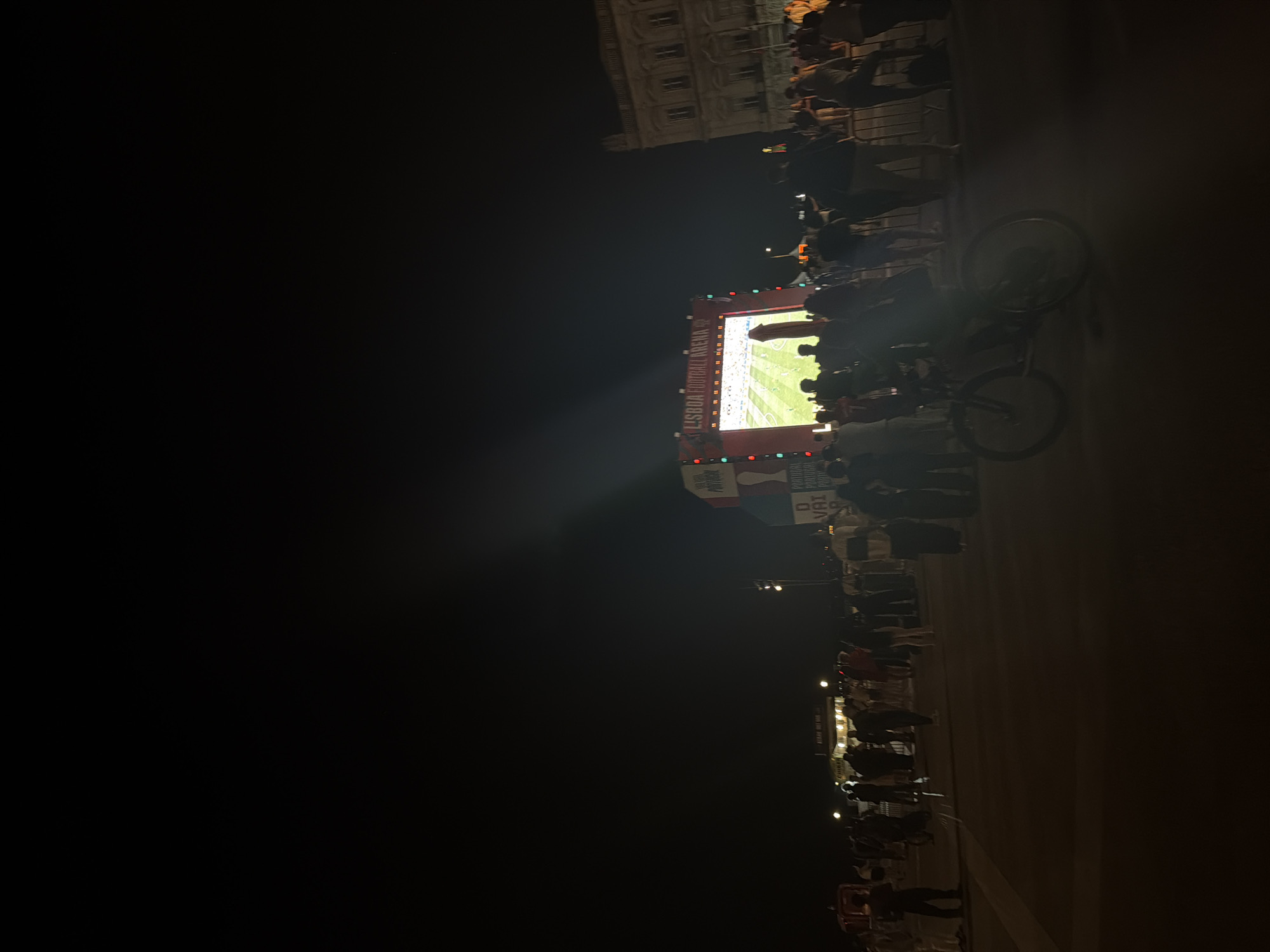 An outdoor nighttime scene shows a large crowd gathered in front of a massive screen displaying a soccer match. The screen, labeled "LISBOA FOOTBALL ARENA," is illuminated and shows players on a green field. People are standing, some are silhouetted against the bright screen, and a bicycle is visible in the foreground. The surrounding environment is dark, with some buildings and streetlights in the background. This event is likely taking place in a public square or plaza in Lisbon, Portugal, possibly related to a major sporting event like the World Cup or European Championship.