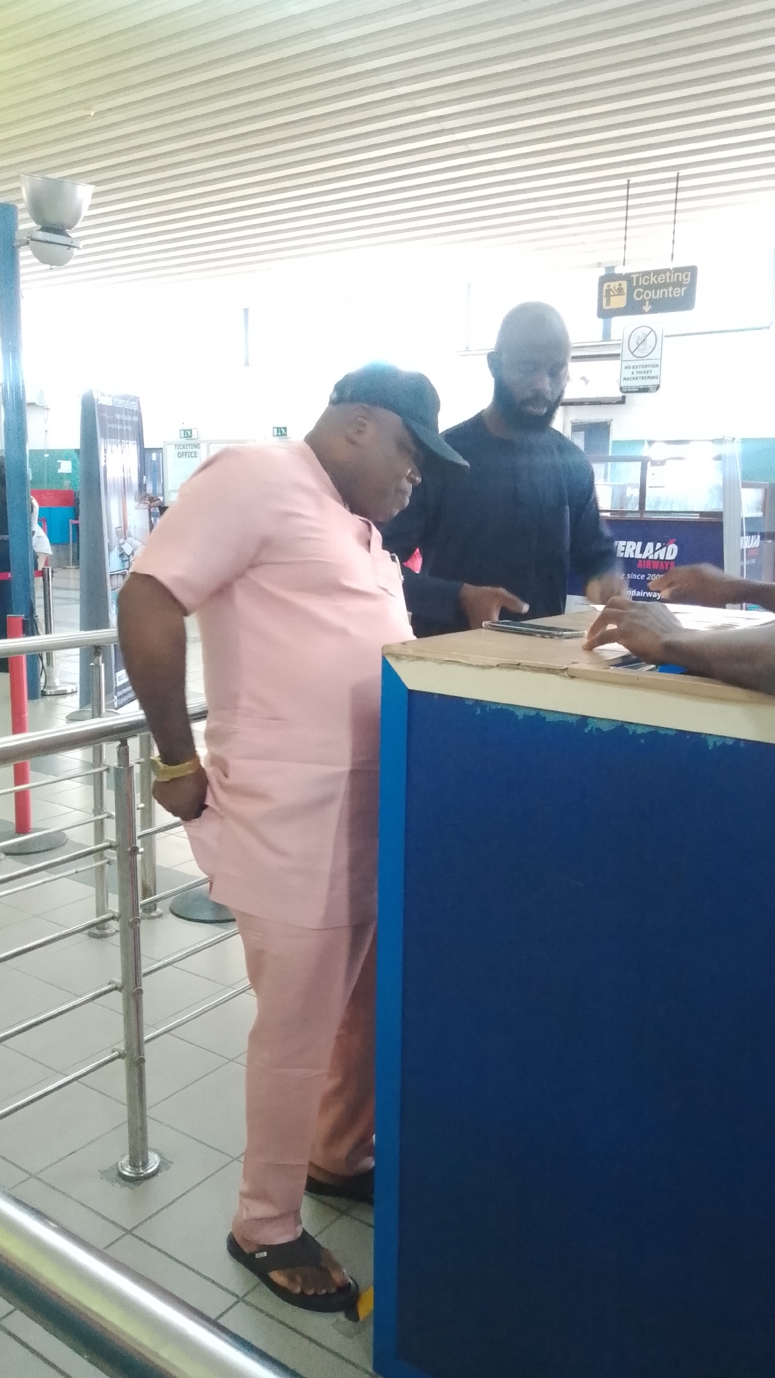 The image depicts two men interacting at a counter, likely in a ticketing office or check-in area, within an airport or transportation hub. The man in the foreground is wearing a light pink traditional Nigerian outfit and a dark baseball cap. He has a gold bracelet on his left wrist and is wearing black flip-flops. The man behind him is wearing a dark long-sleeved shirt and has a beard. They appear to be engaged in a transaction or discussion.
In the background, a sign reads "TICKETING OFFICE" with an arrow pointing down. Another sign below it says "NO EXTORTION TICKET RACKET RING". Part of a flight information board is visible with "ERLAND AIRWAYS" and text below it. The environment appears to be indoors with tiled flooring, metal railings, and a suspended ceiling with horizontal panels. The lighting is bright, suggesting daylight or strong artificial lighting. Based on the context of an airport and signage, this scene could be located at the ticketing counter of an airport in Orerokpe, Nigeria, or a similar transportation facility.