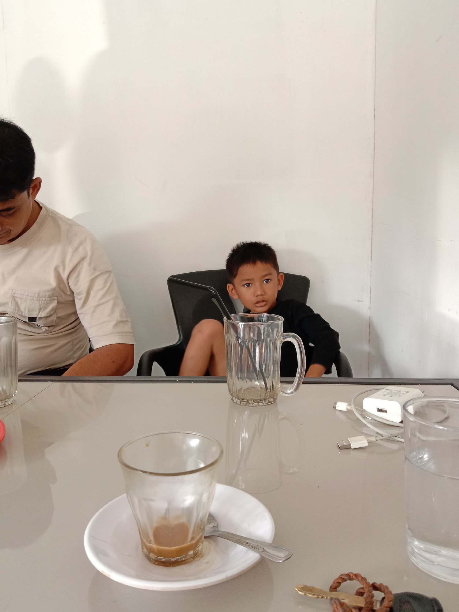 A young boy with short black hair, wearing a long-sleeved black shirt, sits in a black chair at a glossy, light-colored table. He is looking to the right of the frame. Next to him, to the left, is the back of a man with short black hair wearing a tan t-shirt. On the table are several glasses: one empty, one with coffee remnants on a saucer with a spoon, one filled with water, and one with a straw inside. There is also a white phone charger and a set of keys on the table. The background is a plain white wall. The image was taken in Lhokseumawe, Indonesia.