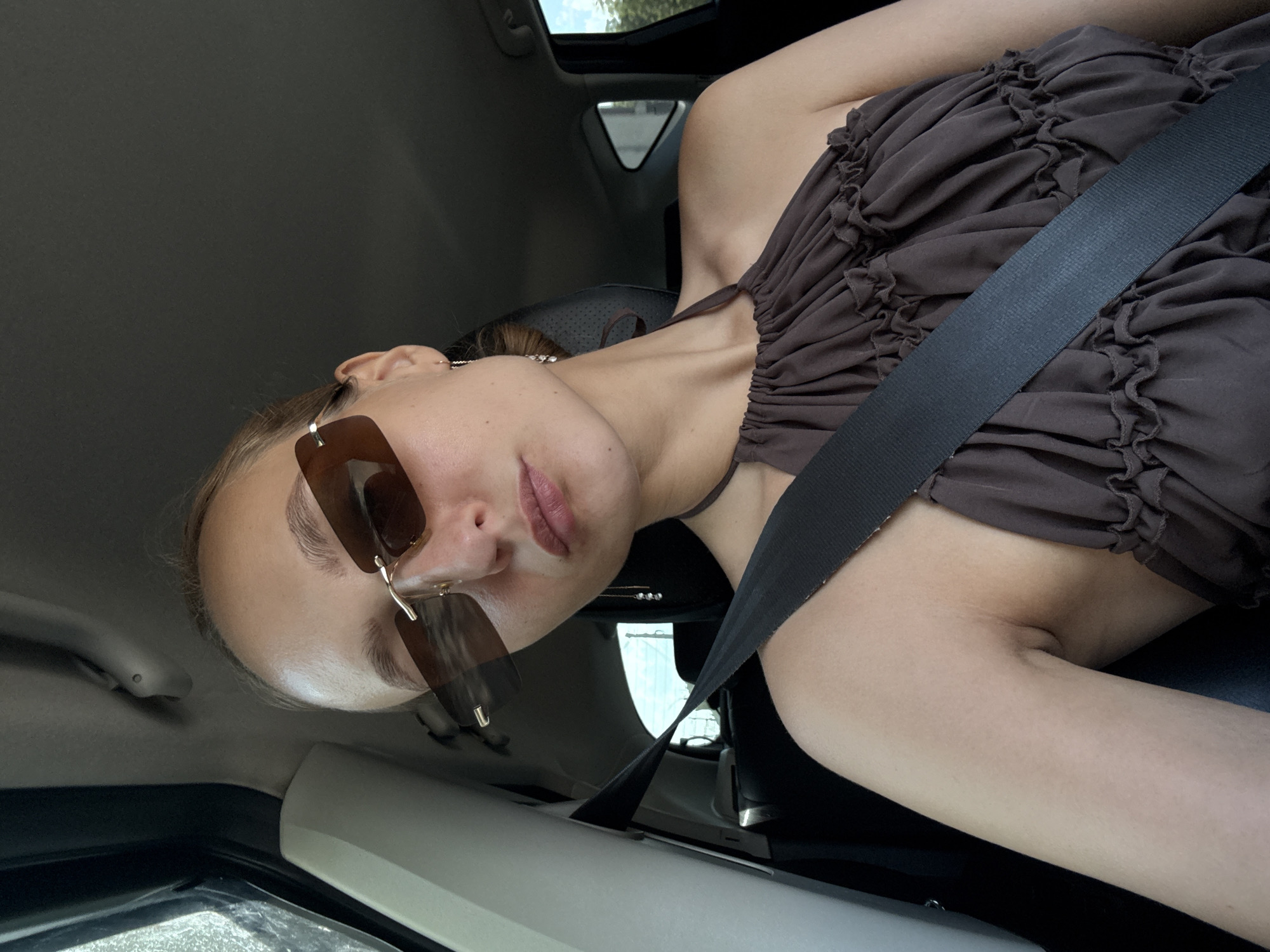 A young woman with light skin and blonde hair pulled back in a bun is sitting in the passenger seat of a car. She is wearing a brown halter top with ruffled detailing and brown-tinted rectangular sunglasses. A black seatbelt is diagonally across her body. The interior of the car is visible, including the headrest and a portion of the driver's seat. The background outside the car shows greenery and a bright sky, suggesting it is daytime. The image was taken in Izhevsk, Russia.