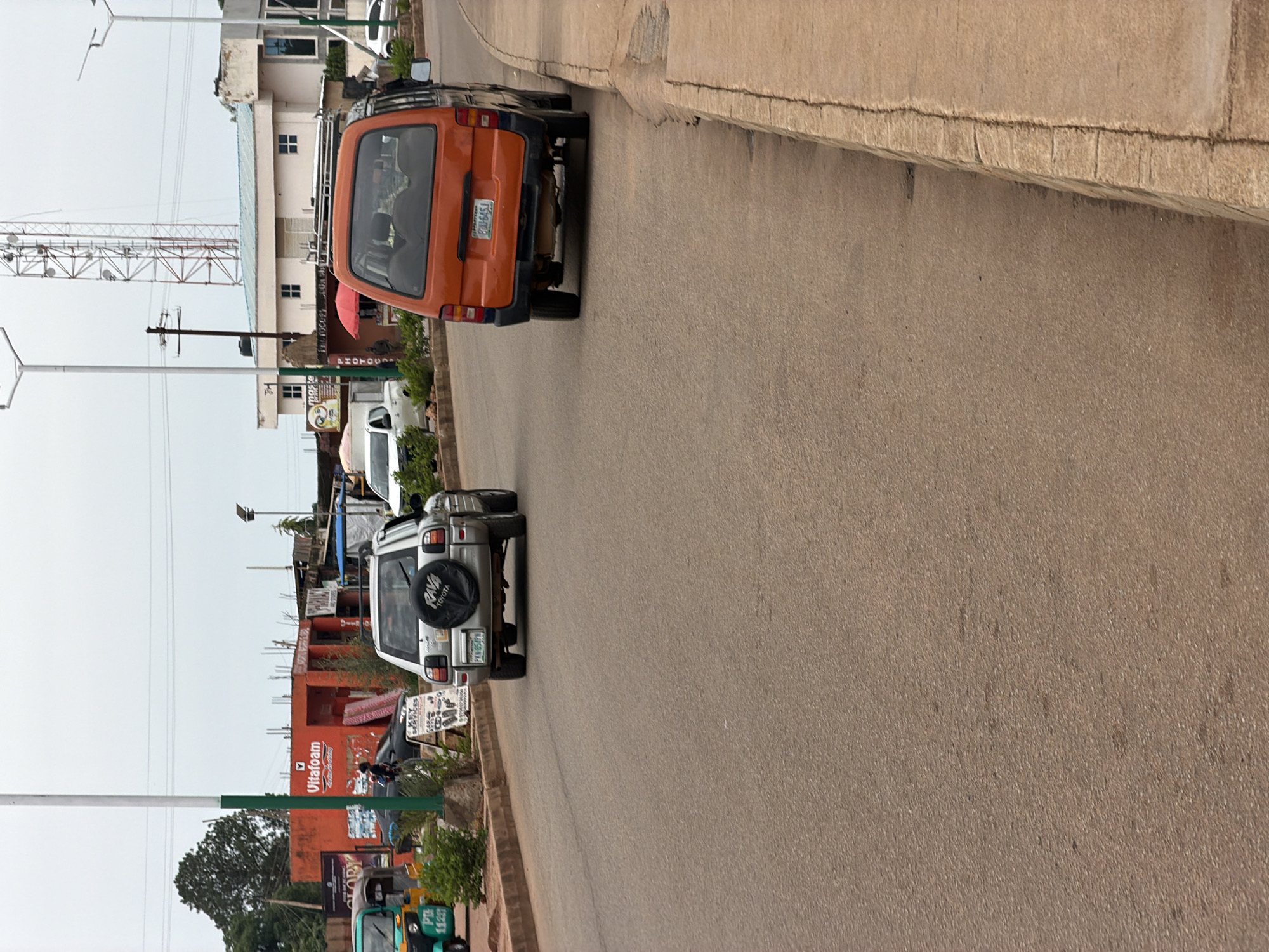 In Bukuru, Nigeria, a silver Toyota RAV4 SUV is parked on the side of a paved road. The SUV has a spare tire mounted on its rear, with a black cover. A license plate with the characters "PKN 854FN" is visible. To the right of the RAV4, a bright orange van is also parked on the side of the road. Its license plate reads "EBU 645J". In the background, several commercial buildings line the street, including one with an orange facade that displays the "Vitafoam" logo. A tall telecommunications tower stands in the distance, and a green and white utility pole is positioned near the foreground. The sky is overcast, suggesting a cloudy day. The road surface is asphalt, and a raised concrete curb runs along the right side of the road. Greenery, including bushes and trees, is visible along the roadside.