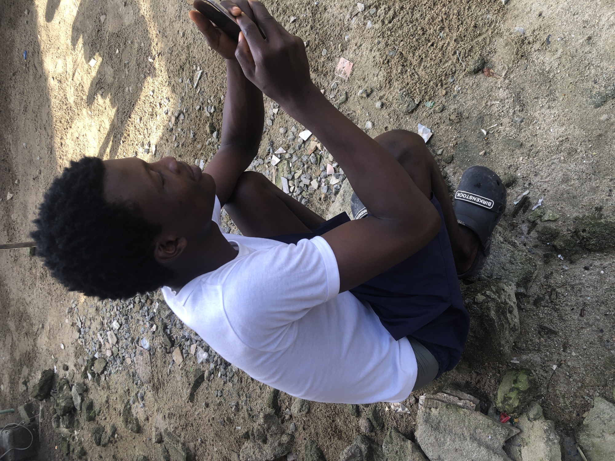 A young Black male wearing a white t-shirt and dark blue shorts is squatting on sandy, gravelly ground in Ajah, Nigeria. He is holding a smartphone in his hands and appears to be looking at it. The background consists of dirt and scattered rocks, with some debris visible on the ground. The lighting suggests it is daytime. He is wearing black slip-on shoes with the word "BIRKENSTOCK" printed in white.
