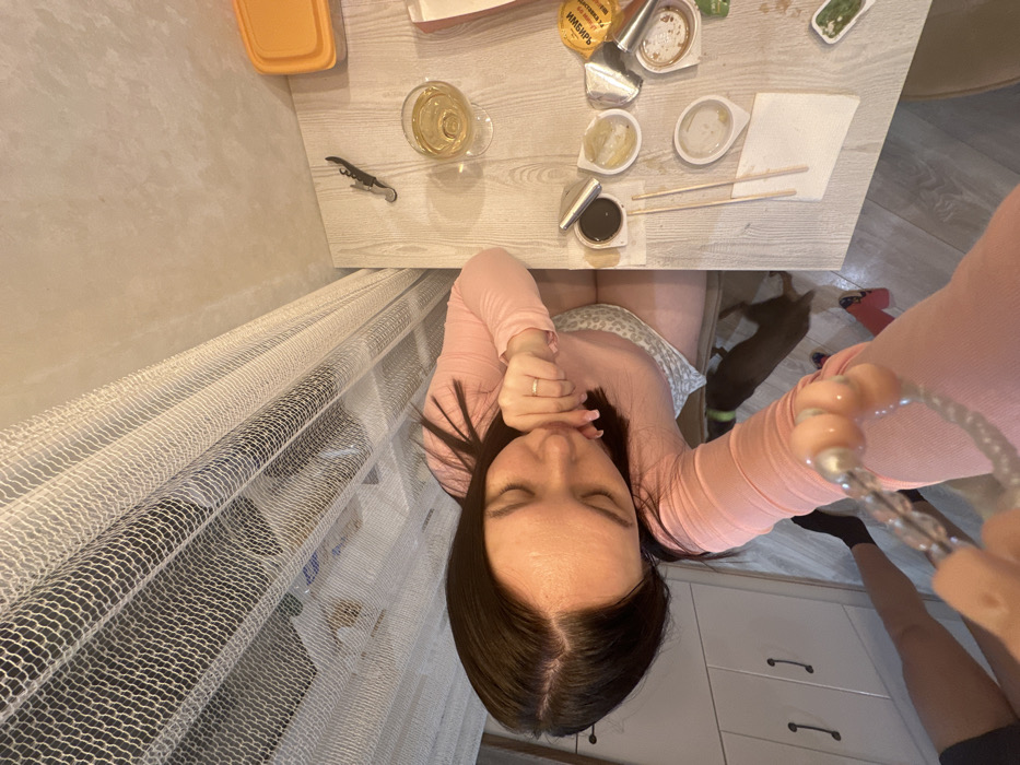 The image captures a high-angle, overhead view of a woman seated at a table, surrounded by food items and household elements.
The central figure is a woman with long, dark hair, parted in the middle. She is wearing a pink long-sleeved top and patterned shorts or a skirt. Her eyes are closed or directed downwards, and one hand, adorned with a simple ring, is held near her chin. Her posture appears relaxed.
To her right, a light-colored wooden table is cluttered with items, suggesting a recent meal. These include a glass containing a light, yellowish liquid, possibly white wine, and a corkscrew. Several small, disposable plastic containers, typical for takeout food, are scattered across the table; some are empty, while others contain remnants of food like soy sauce, wasabi, and ginger. A metallic packet clearly labeled "ИМБИРЬ" (Ginger in Russian) is visible. A pair of chopsticks rests on a white paper napkin. An orange plastic food container is also on the table.
In the background to the woman's left, a window is covered by sheer, white-netted curtains. Below the table, a small, dark-furred dog wearing a light-colored collar is visible. Further down and to the right, a portion of another person's legs, wearing dark socks and possibly shorts or a skirt, can be seen. This person appears to be holding a braided strap with prominent pinkish-clear beads, which might be a phone wrist strap, suggesting they are the one taking the photograph, possibly the woman herself holding the phone.
The setting appears to be an indoor space, likely a kitchen or dining area, with what looks like white kitchen cabinets visible in the lower right corner. The lighting in the image suggests an indoor environment, possibly during the evening or late afternoon due to the warm tone. The overall scene conveys a casual, domestic moment, perhaps enjoying a takeout meal.