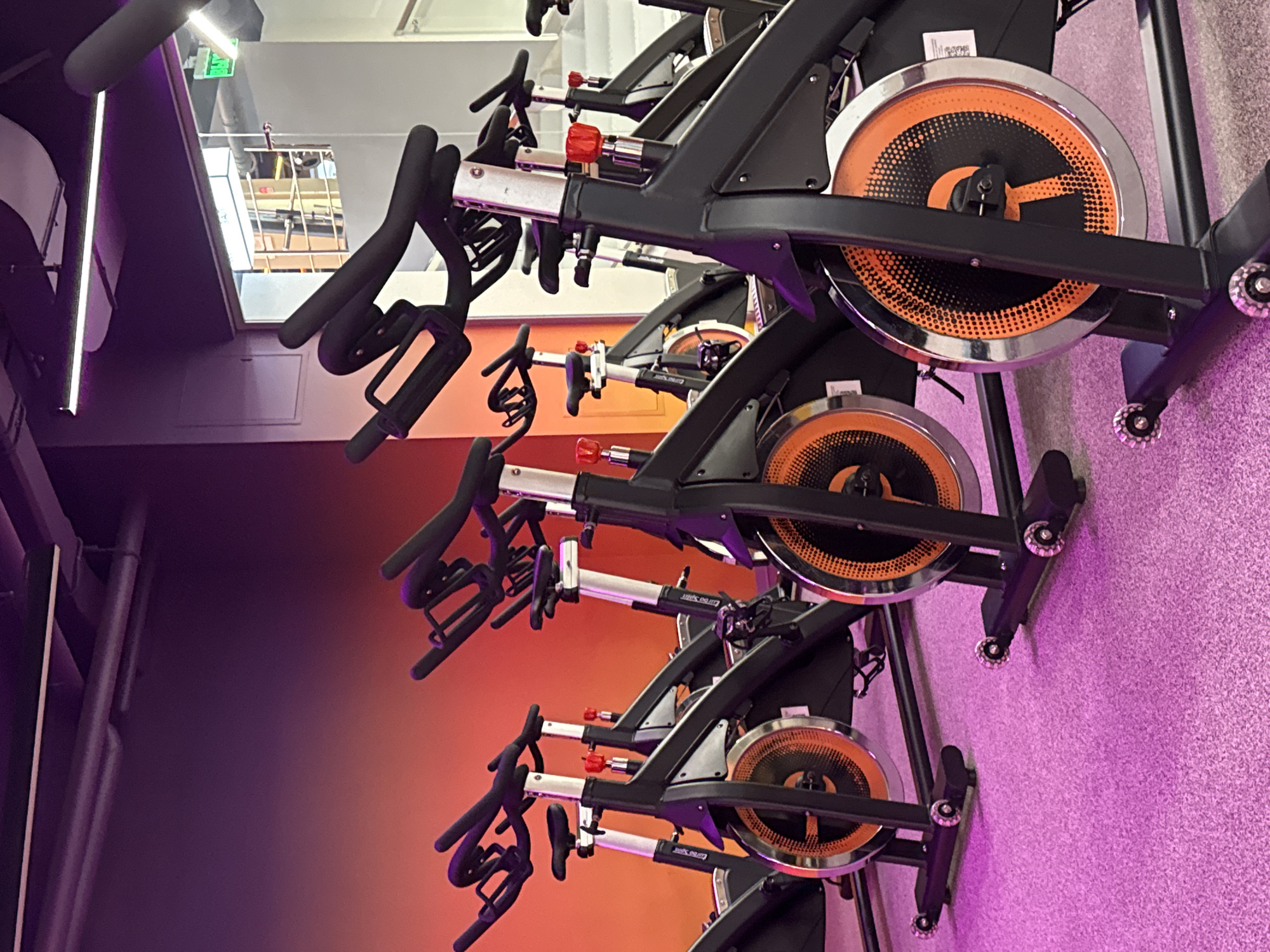 The image shows a row of spin bikes in a gym. The bikes are black with orange detailing on the flywheel. The gym has a color-gradient wall that fades from purple to orange. The floor is covered in a light purple carpet. The exact location in Saint Petersburg, Russia cannot be determined.