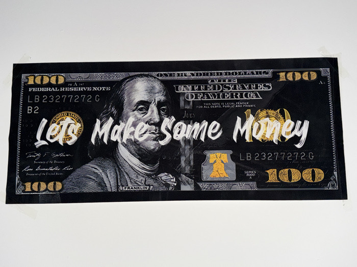 A large, rectangular print, designed to resemble a stylized U.S. one-hundred dollar bill, is affixed to a plain white wall. The artwork features a predominantly black background with white and gold accents. A grayscale portrait of Benjamin Franklin is centered on the left half. Overlaid in white, brush-stroke style text across the center are the words "Let's Make Some Money," with the "O" in "Money" replaced by a gold circular seal resembling the Federal Reserve Seal. Other gold elements include the number "100" in the bottom right corner, a gold image of the Liberty Bell in the bottom center-left, and a gold eagle motif in the upper right. Standard bill details such as "FEDERAL RESERVE NOTE," "UNITED STATES OF AMERICA," "ONE HUNDRED DOLLARS," the serial number "LB 23277272 G," and signatures are depicted. The print is secured to the wall with small pieces of translucent tape visible in the top left and right corners. The scene is located in Amber Valley, United Kingdom.