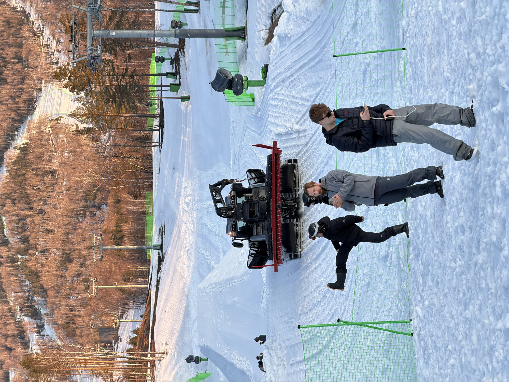 The image depicts a winter scene, likely at a ski resort in Озёрное, Russia. A snow groomer, or snowcat, is positioned in the center of a snowy slope. Three individuals are present: one is looking at his phone, another is looking at the groomer, and the last is bent over. The environment includes a forested area, ski lift infrastructure, green snow fencing, and a snow-covered landscape.