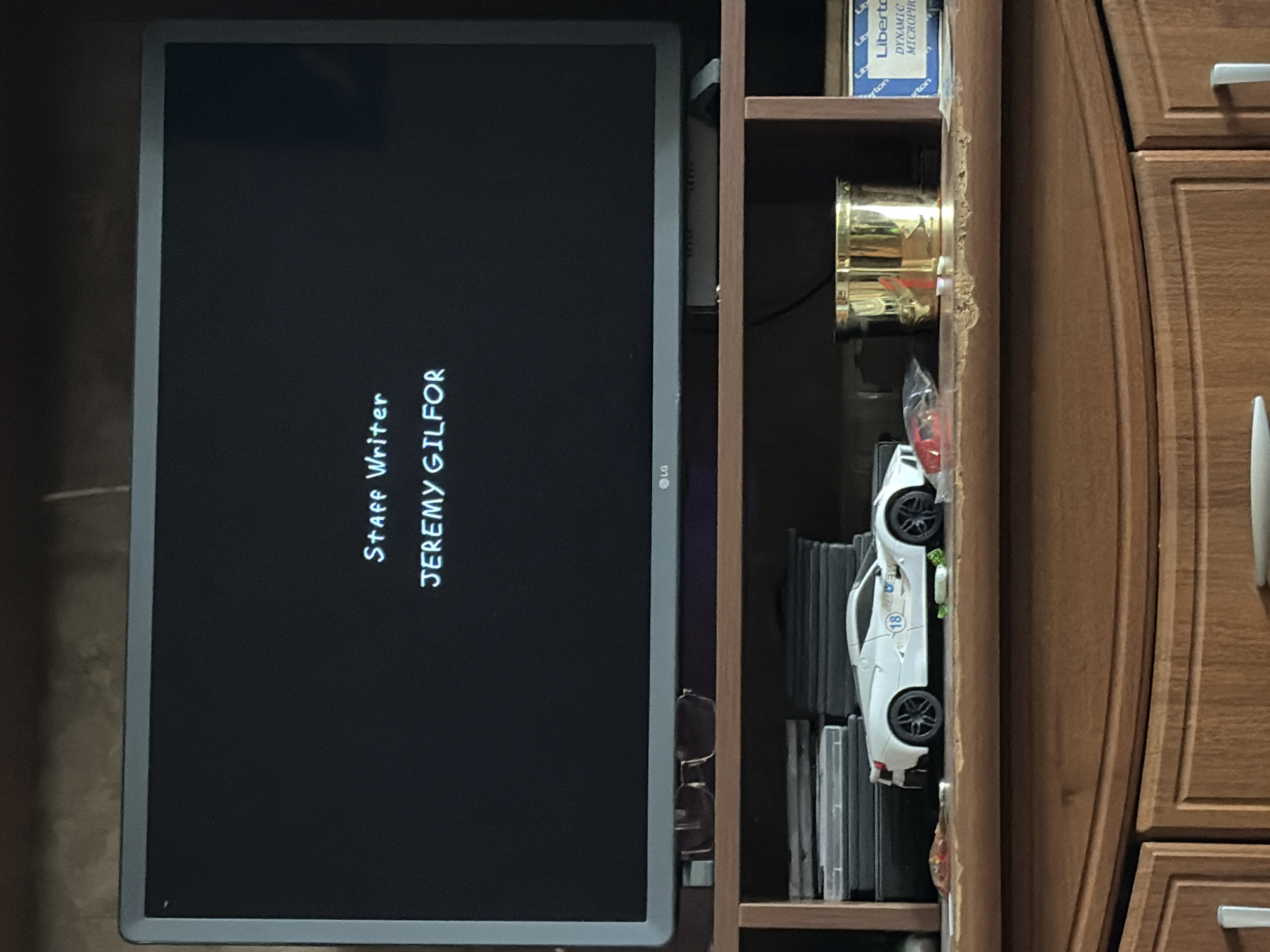 The image shows a television screen displaying "Staff Writer JEREMY GILFOR". Below the screen, a shelf contains several items, including stacks of DVDs or cases, a white toy sports car with the number 18 on its side, a golden cup, and a blue box labeled "Liberto DYNAMIC MICROPH". The television is an LG model. The shelf is part of a wooden entertainment unit. The scene appears to be indoors, in a residential setting.