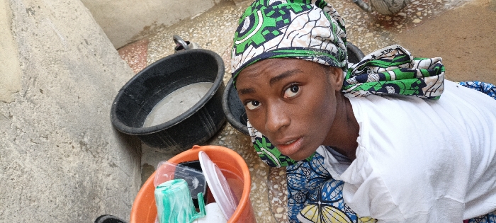 A woman with a green and white patterned headwrap and a white shirt looks up at the camera. An orange bucket filled with various dishes and a green cup sits below her. Behind her, there are two black buckets, one of which appears to contain a gray substance. A concrete wall is visible to the left. Her expression is neutral, with a slight upward gaze.