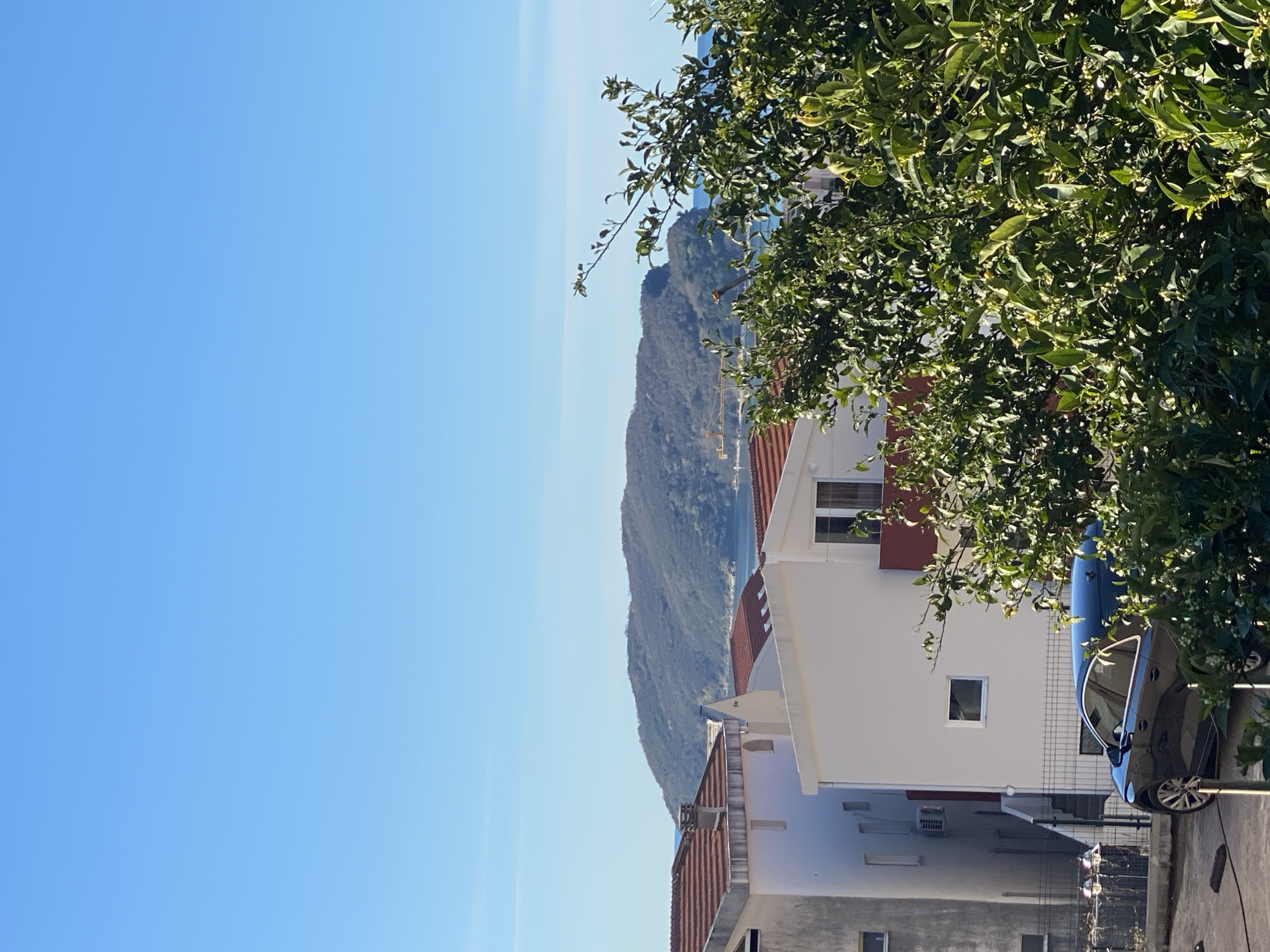 The image is a daytime, outdoor shot in what appears to be Budva, Montenegro. A coastal mountain is visible in the background, with the blue of the Adriatic Sea and the clear blue sky above. In the foreground, the corner of a multi-story white building with a red tile roof is partially obscured by green foliage from a tree. A dark-colored car is parked next to the building. The location is likely a residential area with a view of the surrounding mountainous landscape and the sea.