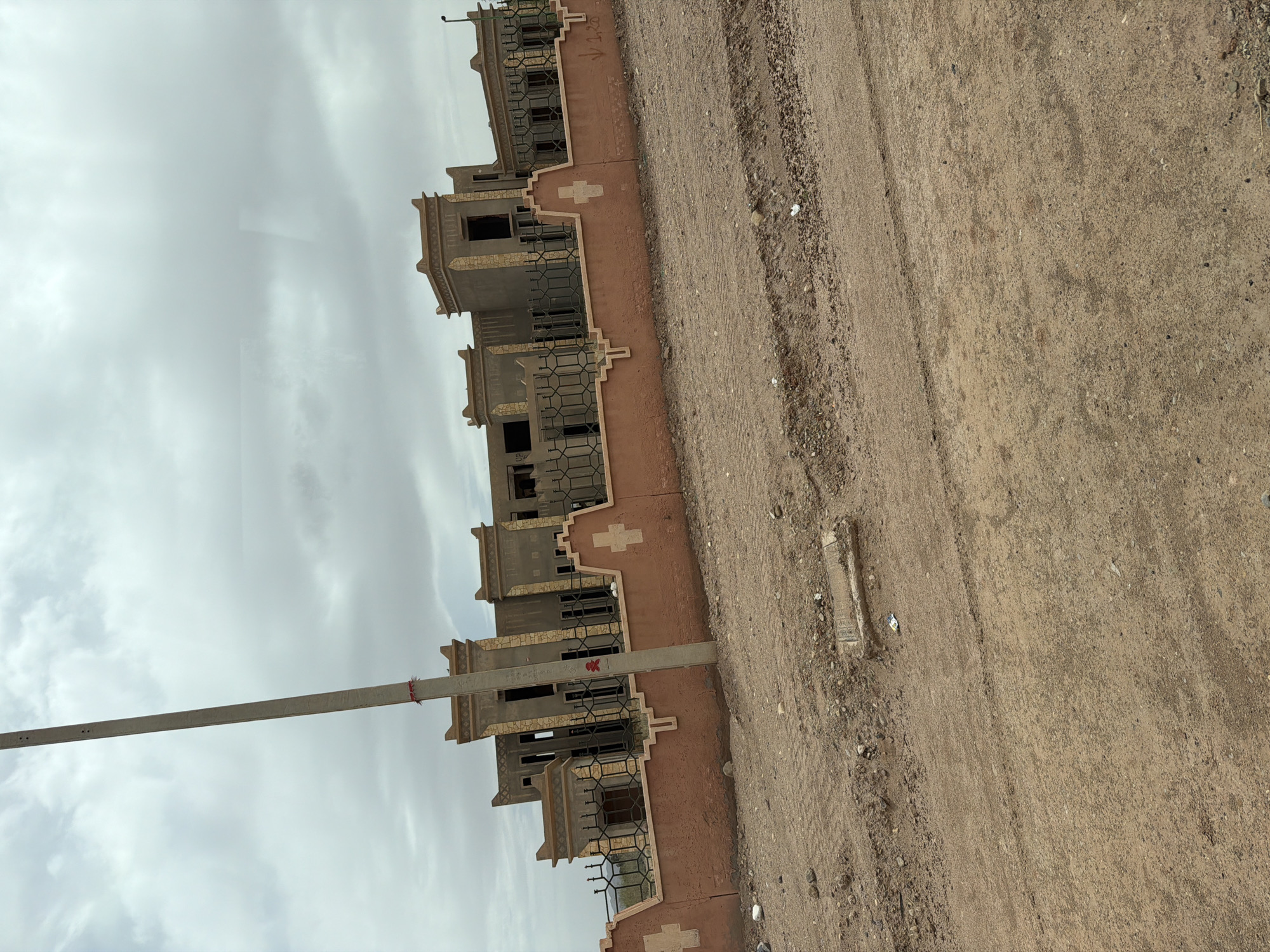 The image shows a row of unfinished buildings in Tarmigt, Morocco. The buildings are constructed with a beige or light brown concrete base, featuring architectural details like window frames and decorative crosses. Some windows have black metal grilles. A concrete utility pole extends vertically from the bottom to the top left of the frame. The sky is overcast. The ground is dry, dusty dirt, showing tire tracks and minor debris. There are no visible people in the scene.
