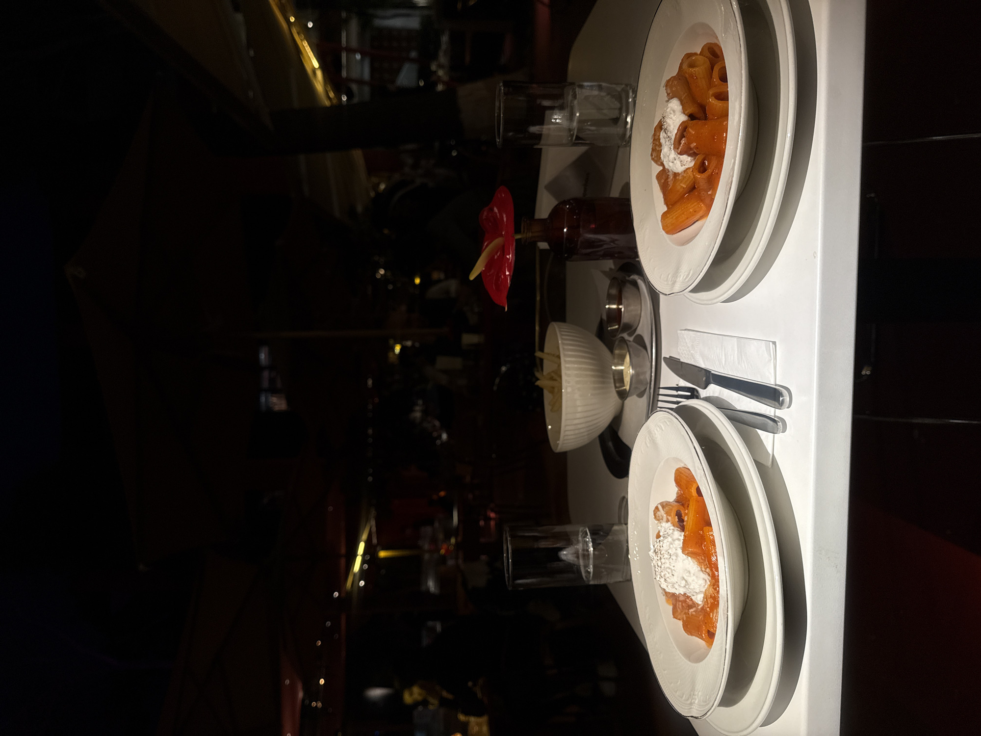The image captures a dining table in what appears to be a restaurant setting, likely in Yerevan, Armenia, given the cultural references often associated with the city. The table is set for two, featuring two plates of pasta with a tomato-based sauce and a dollop of cheese on top. Cutlery is neatly arranged beside each plate, along with glasses filled with liquid. A red anthurium flower in a small bottle serves as a decorative centerpiece. A small white bowl is also visible, containing an indeterminate substance. The lighting is dim, suggesting an evening meal or a dimly lit interior ambiance. Background elements hint at a restaurant interior, though details are obscured by the darkness.