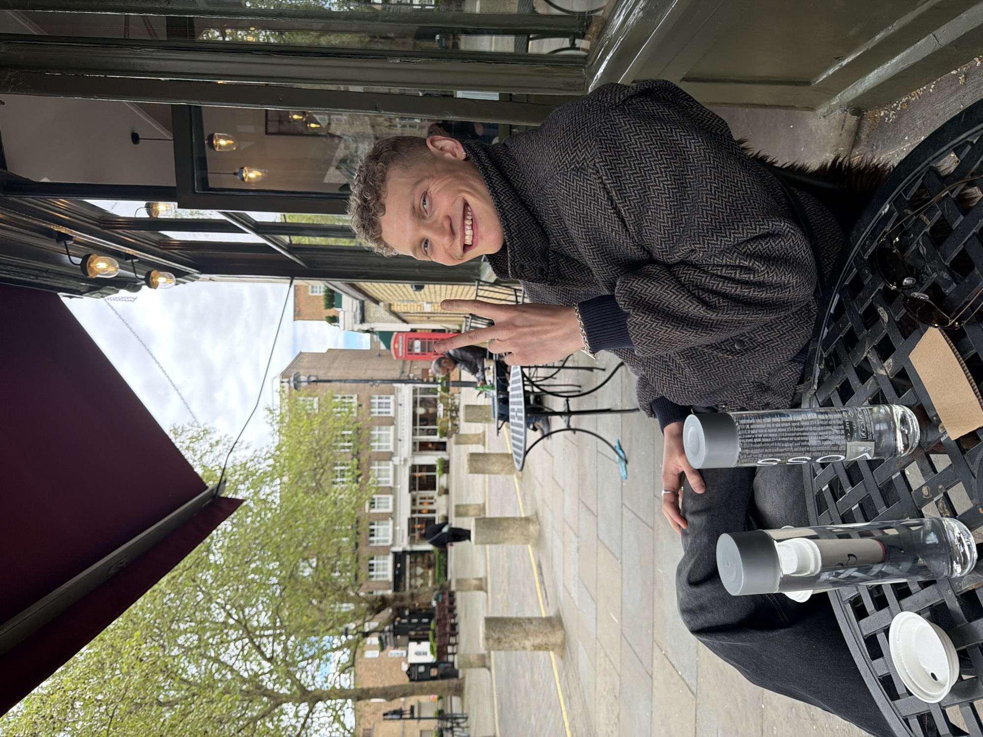 The image captures a smiling young man seated at an outdoor black metal table, giving a peace sign with his left hand towards the camera. He has light brown, curly hair and a wide, joyful smile, revealing his teeth. He wears a dark, herringbone-patterned jacket, possibly tweed, over a dark long-sleeved top, and has a small earring in his left ear and a silver bracelet on his right wrist.
On the table in front of him are two clear glass water bottles with grey tops; one displays the word "GOOD" and smaller text. A white plastic lid and a pair of dark sunglasses also rest on the table. A burgundy awning is partially visible in the upper left of the frame.
The background depicts a charming urban street scene, identifiable as London, United Kingdom, given the distinctive red telephone booth visible behind the man. The street is paved with light-colored flagstones and lined with concrete bollards. Various multi-story buildings with traditional white-framed windows line the square, and fresh, light green leaves on trees suggest a springtime setting. The sky appears overcast, indicating a cloudy daytime. The man's cheerful expression conveys a relaxed and positive mood while enjoying an outdoor moment.