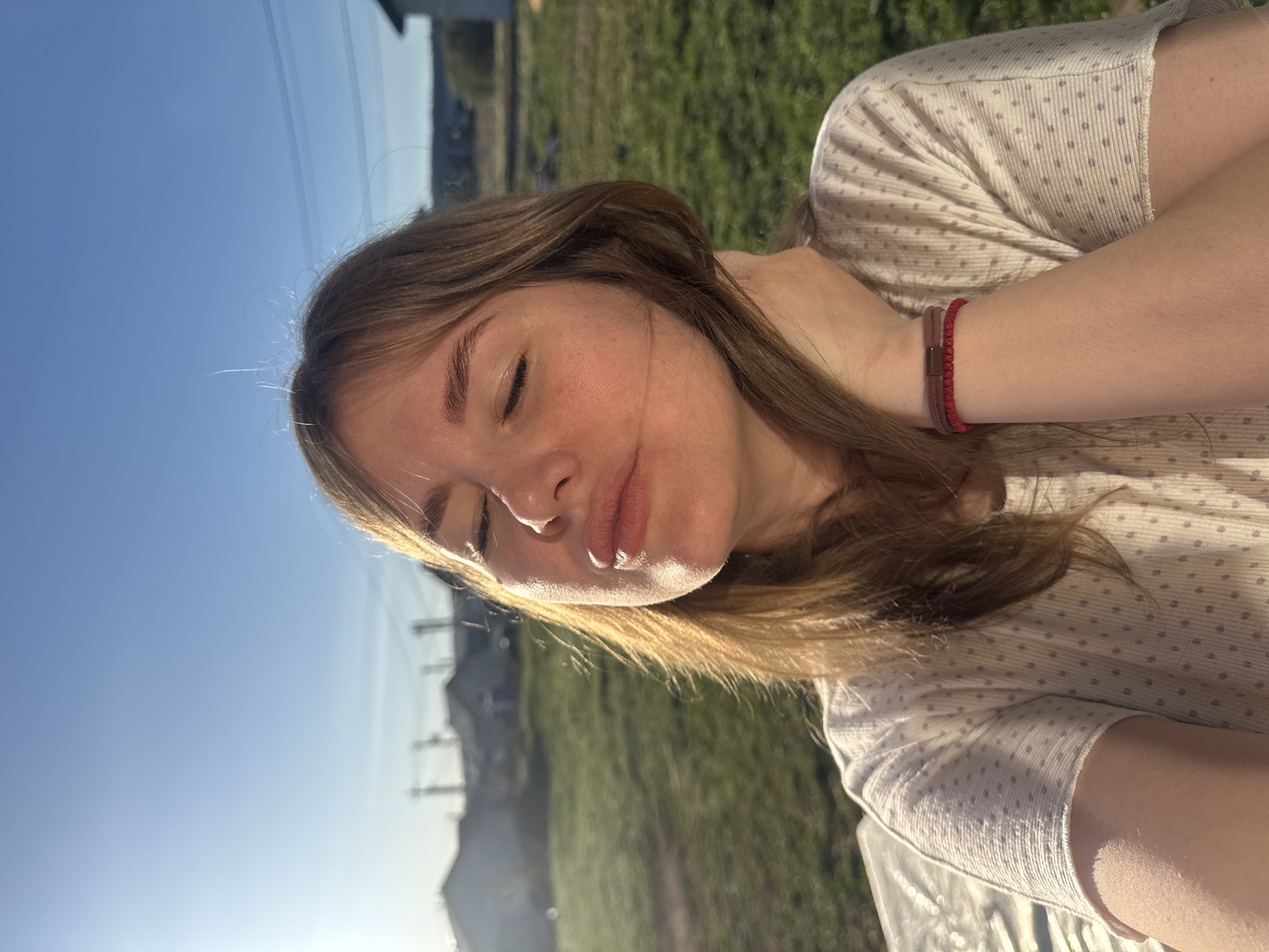 A young, light-skinned woman with closed eyes and light brown hair, wearing a light-colored, polka-dotted short-sleeved shirt and red bracelet on her left wrist, holds her neck with her left hand. The scene takes place outdoors in Ферма, Russia, as seen by the power lines in the background and a grassy field behind her. The sun is shining, casting shadows on her face and the surrounding landscape.