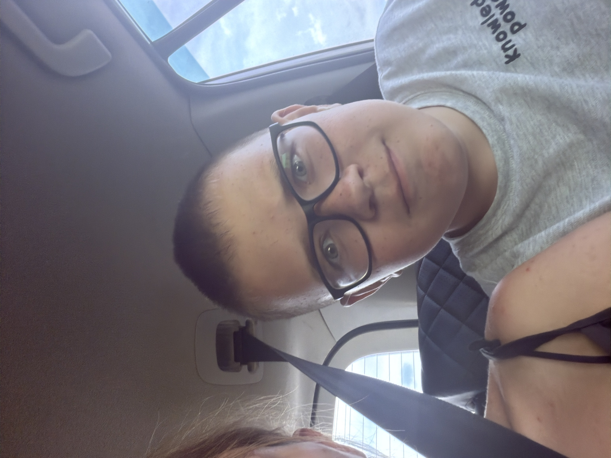 In Ostrovka, Russia, a young boy with short brown hair and glasses is taking a selfie inside a car. He is wearing a grey t-shirt that says "knowledge power". Another person, partially visible on the left, has blonde hair and is also in the car. The interior of the car shows a seatbelt and part of a quilted car seat. The background outside the car window suggests a cloudy sky.