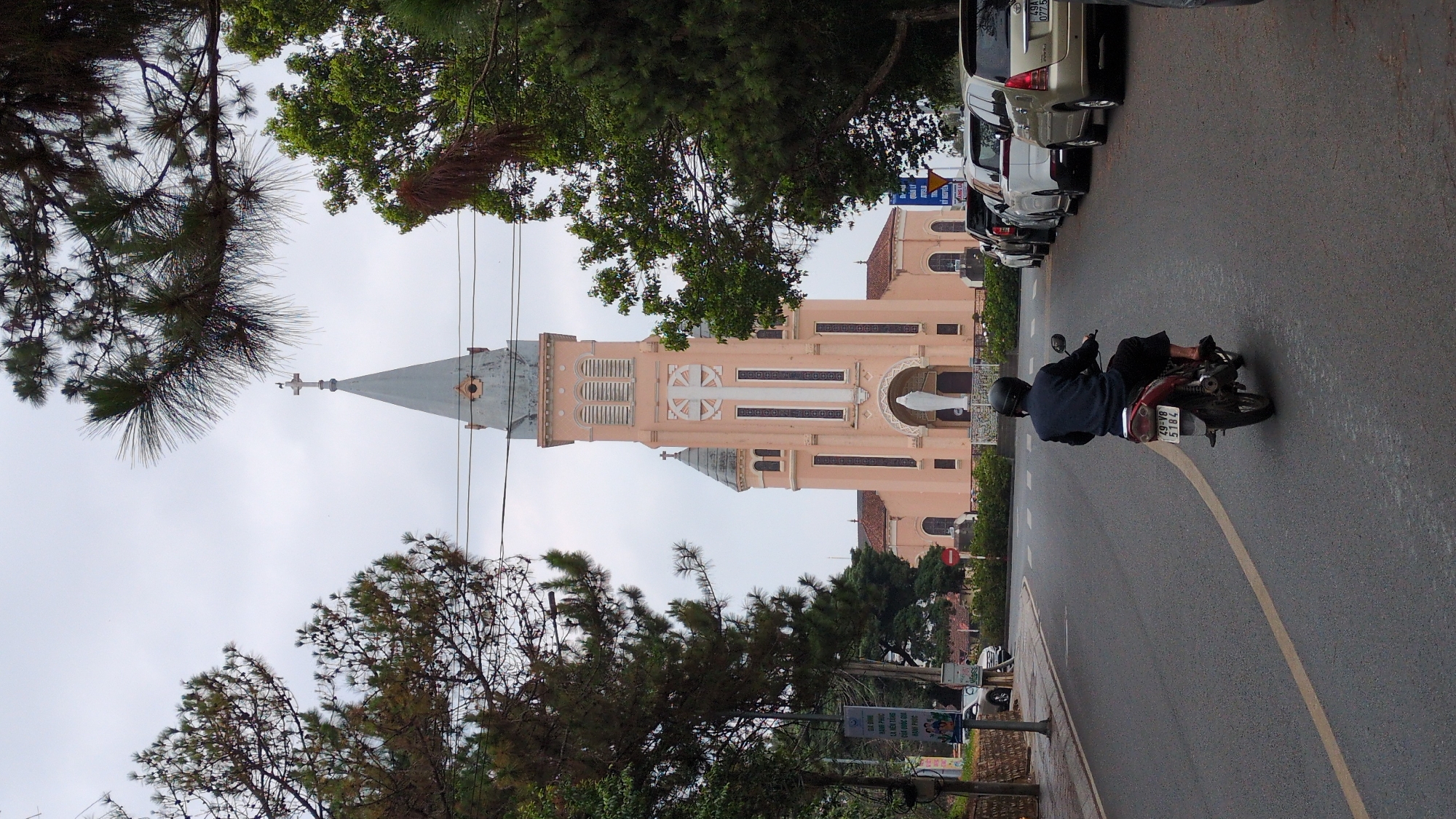 The image depicts a street scene in Da Lat, Vietnam, showcasing the pink Da Lat Cathedral in the background. The sky is overcast. A person wearing a helmet and dark clothing rides a motorcycle on the wet road, in the direction away from the camera. Cars are parked along the side of the road opposite the church. Trees frame the view of the cathedral. A road sign stands near the sidewalk. The photograph is rotated 90 degrees clockwise.