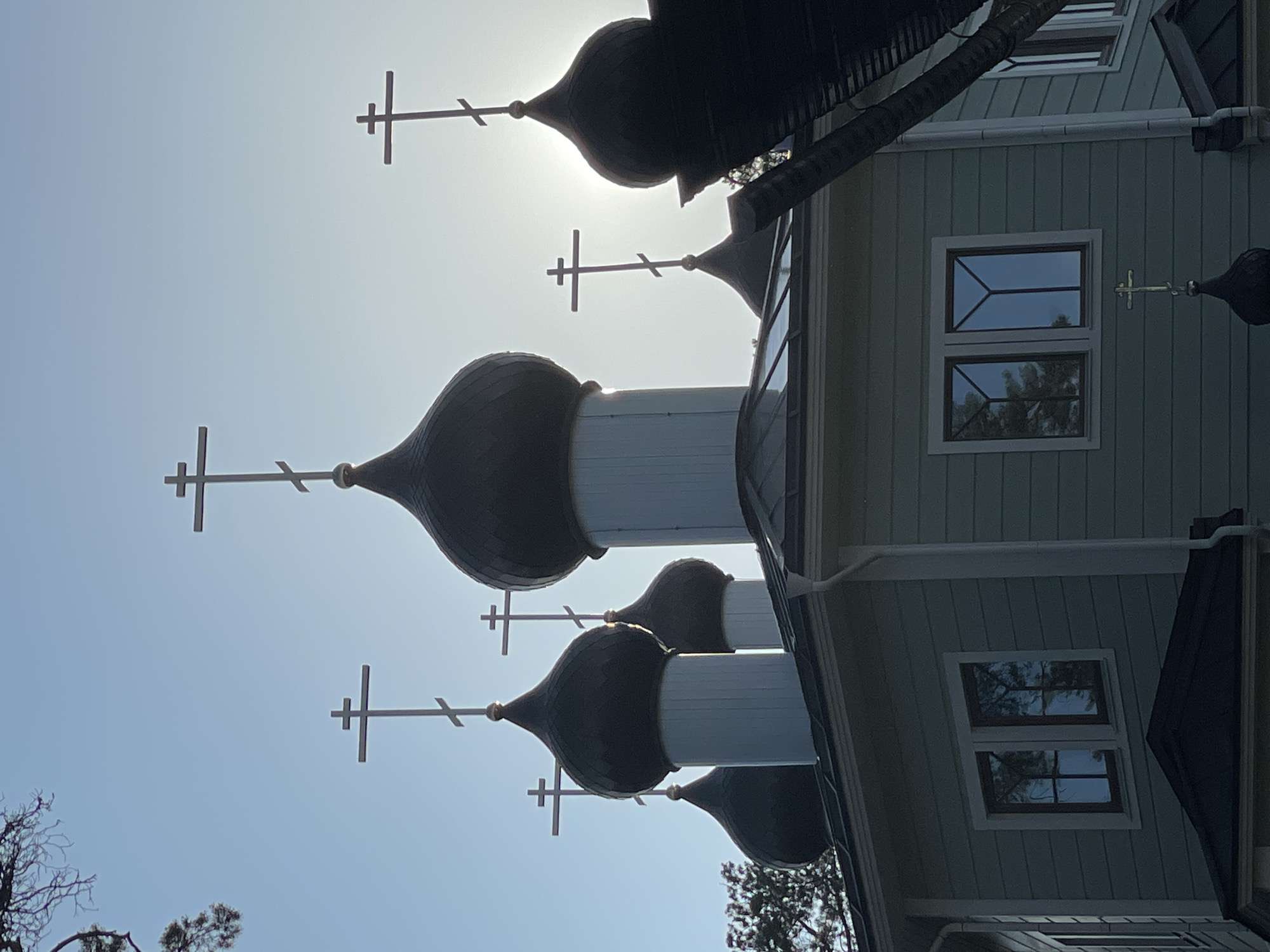 This is a low-angle, outdoor, medium shot of an Orthodox church building with multiple onion domes topped with crosses. The building's exterior is clad in light green siding. Several windows are visible, reflecting the sky and trees. A tree with bare branches is present in the foreground. The sun is bright, creating a lens flare and silhouette effect on some of the domes and crosses. The sky is clear and blue, suggesting a sunny day. The perspective is tilted, with the church and sky dominating the frame. The scene appears to be set in Solnechnoye, Russia. No people or text are visible in the image.