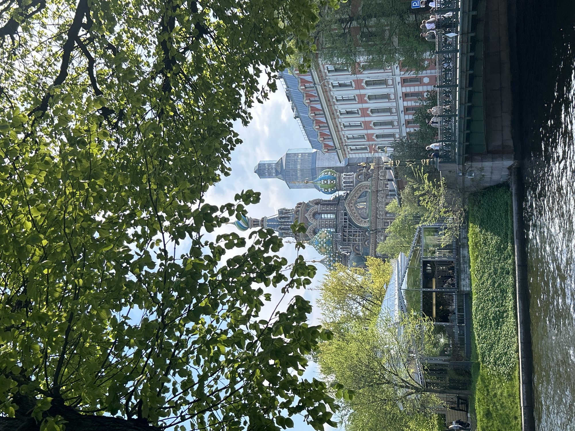 The image shows a view of Saint Petersburg, Russia, featuring the Church of the Savior on Spilled Blood framed by tree branches. The building is adorned with colorful onion domes. A modern glass structure is visible in the foreground, possibly a cafe or greenhouse. A canal runs along the right side of the image. A bridge with people walking on it is visible. Buildings with red and white facades line the canal. In the distance, a modern skyscraper stands out. The sky is partly cloudy.