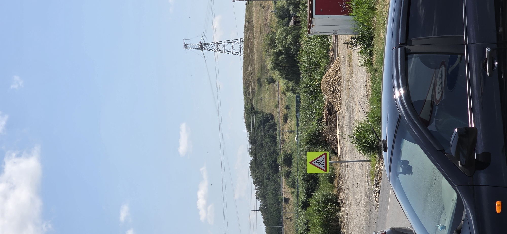 The image depicts a rural or suburban roadside scene in Inozemtsevo, Russia, under a clear blue sky with scattered white clouds. In the foreground, a black car is partially visible, facing left, with its windshield reflecting the surroundings. To the left of the car, another vehicle's rear is visible.
Further into the scene, a yellow pedestrian crossing sign with a red border is mounted on a metal pole. The sign shows a silhouette of a person walking across a crosswalk. Behind the sign, a pile of gravel and construction debris is present, along with dry grass and other vegetation.
In the middle ground, a gentle, grassy hill is covered with trees and brush. A prominent, tall electrical pylon stands on the crest of the hill, with power lines extending across the frame. Beyond the hill, another, smaller electrical pole is visible on the right. A small, weathered wooden structure, possibly a shed or gazebo, can be seen amongst the trees on the right side of the hill. A white and red container or small building is located to the right of the gravel pile.
The overall environment suggests an area undergoing some form of development or maintenance, with a mix of natural landscape and infrastructure.