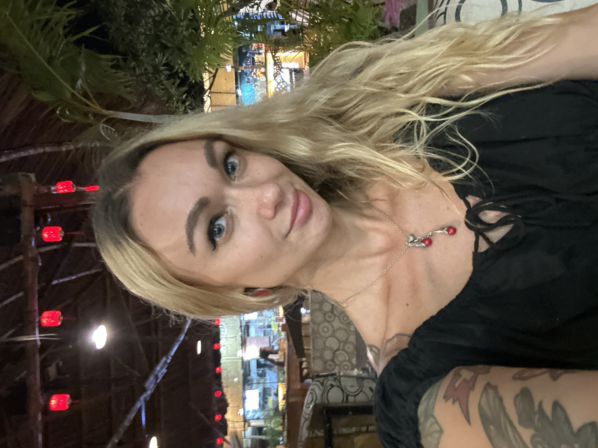 This media file is a selfie of a young woman, captured at night, in what appears to be an open-air restaurant, bar, or resort setting in Phu Thuy Ward, Vietnam. The image is rotated 90 degrees clockwise.
The subject is a woman with long, wavy blonde hair, blue eyes, and light skin. She is looking directly at the camera with a gentle, friendly smile, indicating a relaxed and pleasant mood. She is wearing a black top with a gathered neckline and a silver necklace adorned with a distinctive red, berry-like pendant. A subtle tattoo is visible on her left arm or shoulder.
The background, illuminated by various artificial lights, features lush green foliage, possibly tropical plants or palm fronds, consistent with a warm climate. Prominent red string lights or lanterns are visible on the left side and in the distance, casting a warm glow and suggesting an Asian aesthetic. Other blurry lights are visible further back, hinting at a lively evening environment. Elements of what appears to be a thatched roof structure are also discernible, along with patterned upholstery on furniture, contributing to the ambiance of a casual yet inviting establishment. The overall scene suggests a pleasant evening out. There is no visible text in the image.