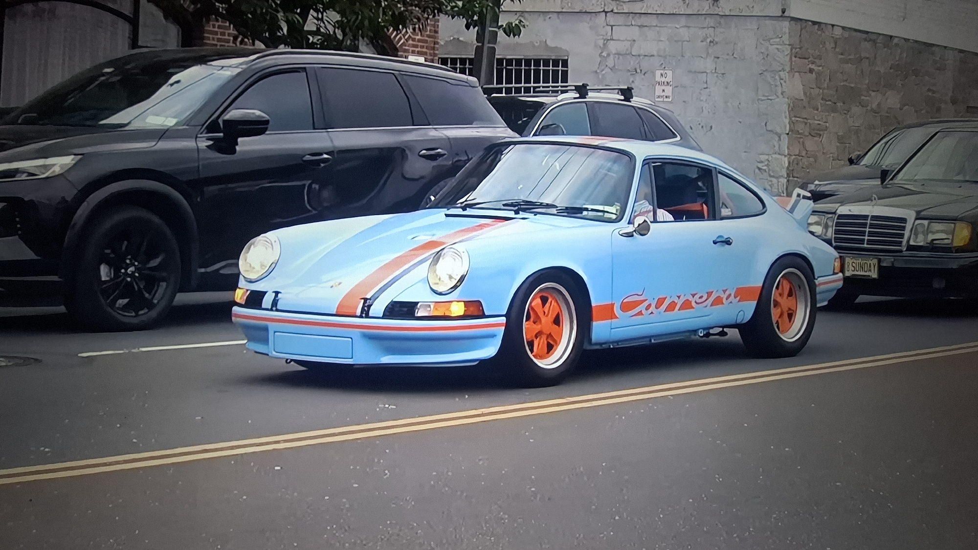 The primary subject of the image is a vintage light blue Porsche 911, likely an early air-cooled model, driving on a paved road during daytime. The car is customized with distinctive orange racing stripes over the hood and roof, and a prominent orange "Carrera" script on its side. It features matching orange wheels, possibly a modern take on Fuchs-style alloys, and a rear "whale tail" spoiler. A person wearing a white shirt is visible in the driver's seat.
The Porsche is positioned in the foreground, with two yellow double lines marking the center of the road. To its left, a dark-colored modern SUV is partially visible. To its right, behind the Porsche, an older model dark Mercedes-Benz sedan is parked or driving, with its front end and grille visible. The Mercedes' license plate clearly shows "8 SUNDAY". Further back, another dark vehicle with a roof rack is partially obscured by the Porsche and an overhead structure.
The scene depicts an urban or suburban street with buildings in the background. One building features a stone facade, and another has brickwork. A white sign affixed to the stone wall reads "NO PARKING DRIVEWAY". The bright lighting indicates it is daytime, and the weather appears clear. The image captures a vibrant classic car in motion within a street setting alongside other vehicles.