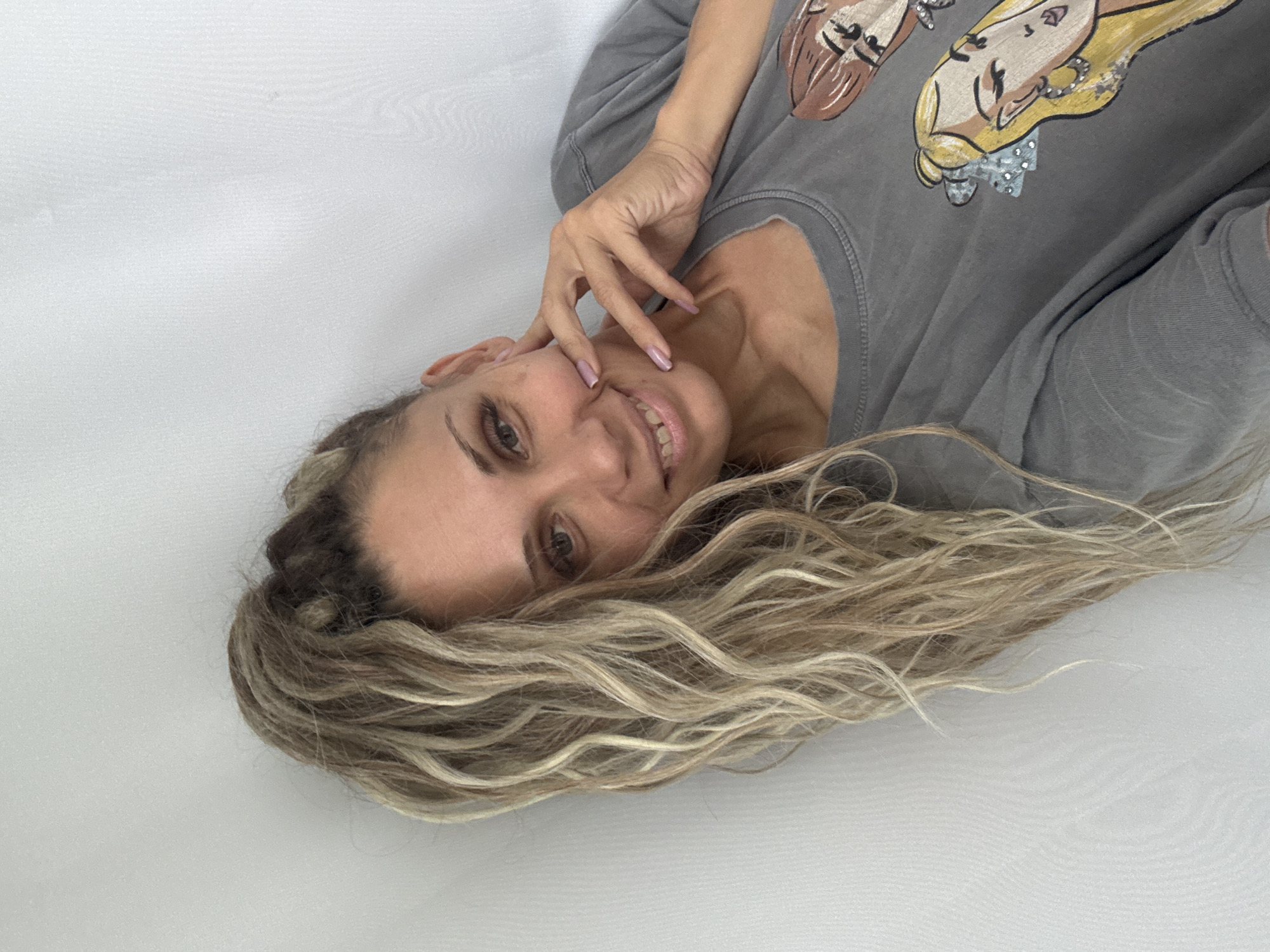The image shows a woman with long, blonde, wavy hair pulled up in a ponytail, leaning against a white wall. She is wearing a gray t-shirt with cartoonish drawings of women on it. Her right hand is resting on her cheek, and she is smiling. There is no visible information or context that would place this image in the city of Dumino, Russia.