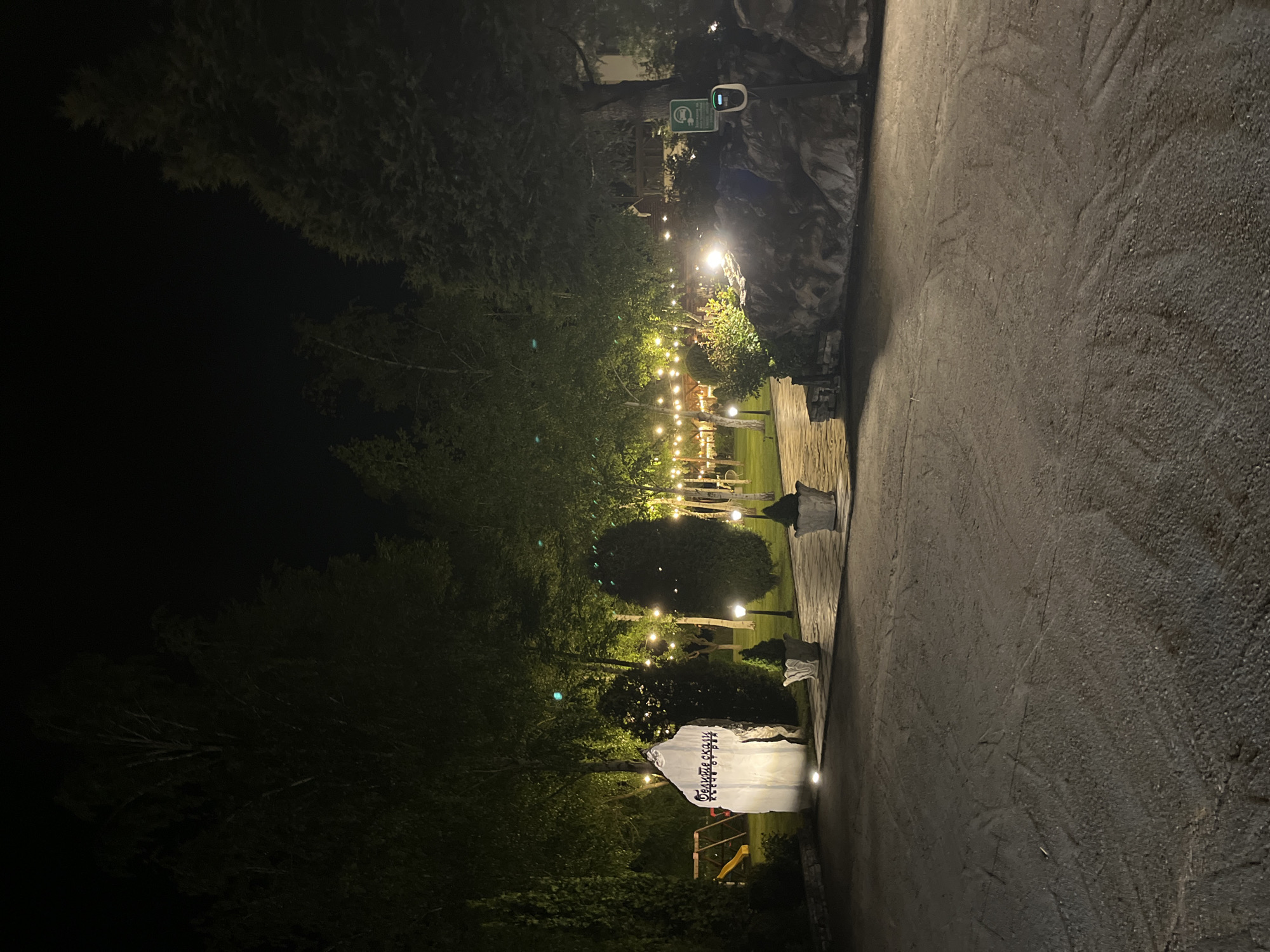 The image shows an outdoor scene at night in Koprivlen, Bulgaria. A paved pathway runs along the right side of the image. To the left of the path, there is a garden with trees and shrubbery illuminated by strings of lights. A structure with writing on it stands at the bottom left of the garden. There are also some stone planters visible along the garden path. A sign is present above the garden area. The atmosphere is dark, but the lights provide illumination to the garden.