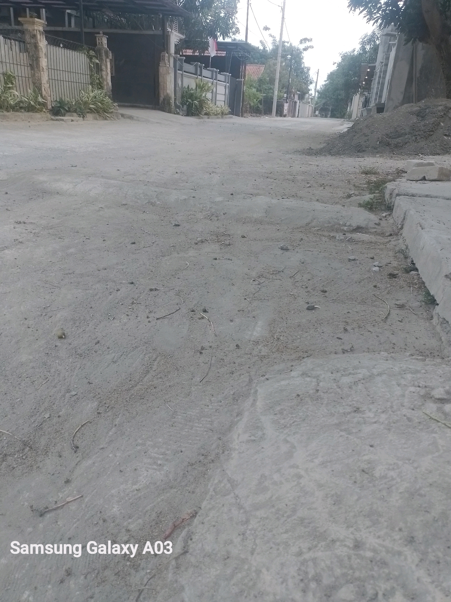 The image depicts a paved street in what appears to be a residential area. The street surface is uneven and made of gray concrete or asphalt. On the left side of the street, there is a fence made of metal bars and stone pillars, with some greenery planted along the fence. Beyond the fence, there are buildings with tiled roofs and an open gate. On the right side of the street, there is a pile of dark soil and some concrete blocks. The street extends into the distance, lined with trees and houses. The sky is overcast. The text "Samsung Galaxy A03" is visible in the bottom left corner of the image. This scene is consistent with typical residential streets found in many Indonesian cities, including Indramayu, with its mixture of housing, infrastructure, and the presence of construction materials.
